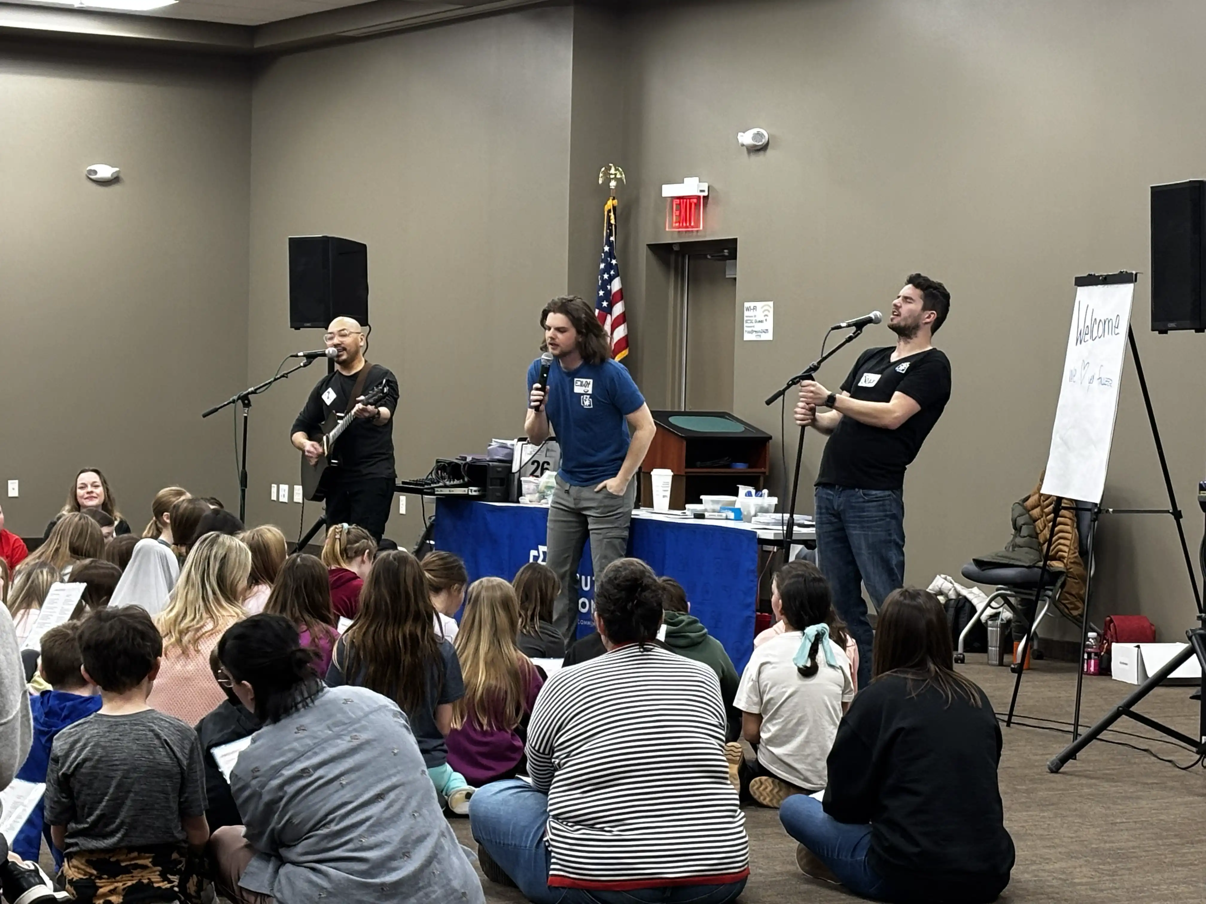 Music presenters singing to a crowd gathered on the floor and sitting in chairs