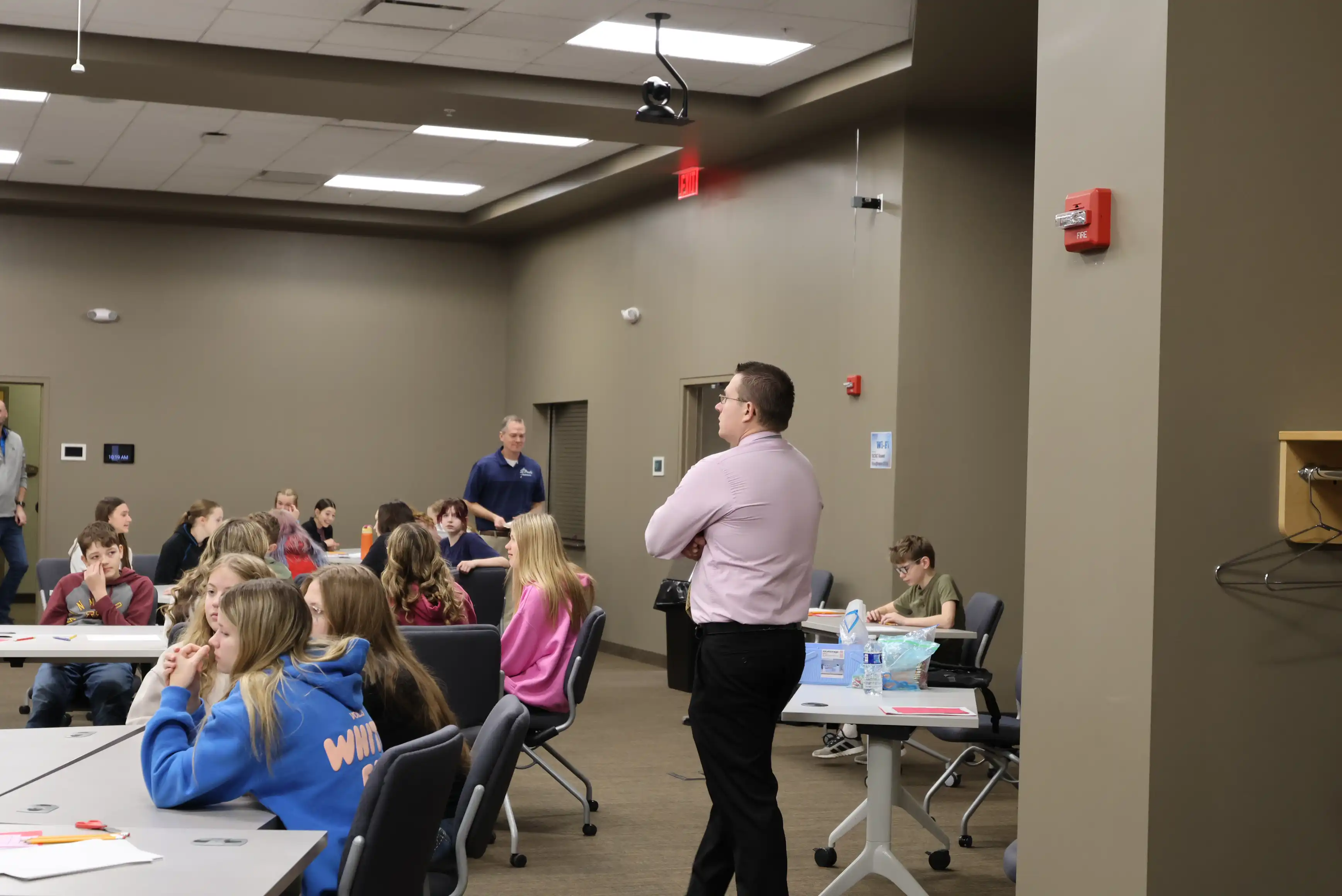 students sitting at tables listening while parents stand around