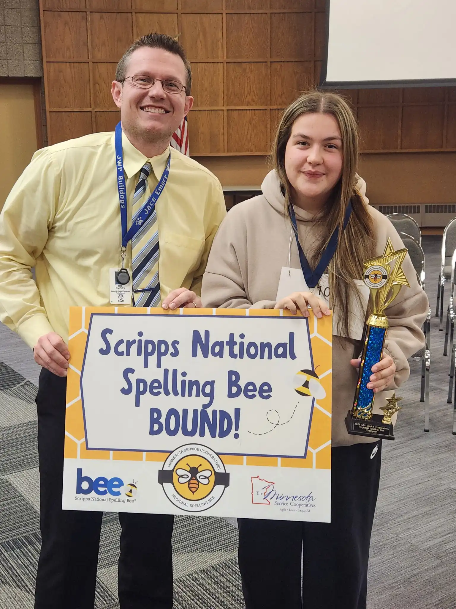 student smiling next to an adult holding a sign that says "Scripps National Spelling Bee Bound"