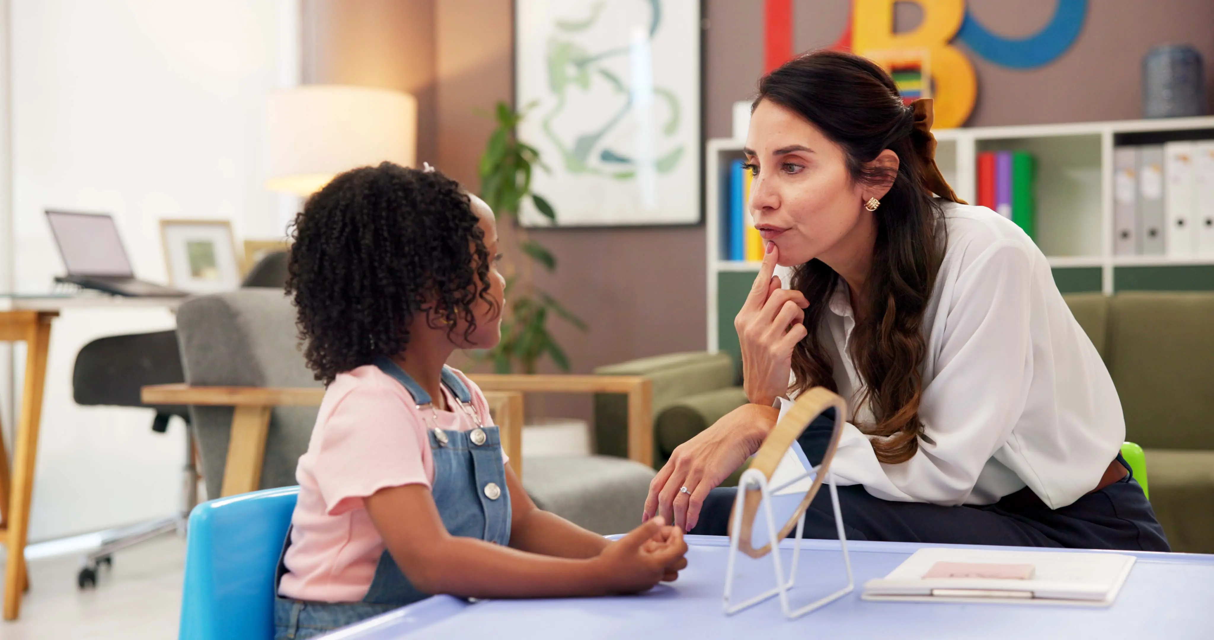 Speech teacher working with student learning how to move their mouth to make certain sounds. There is a mirror and both teacher and student have a finger to their mouth.