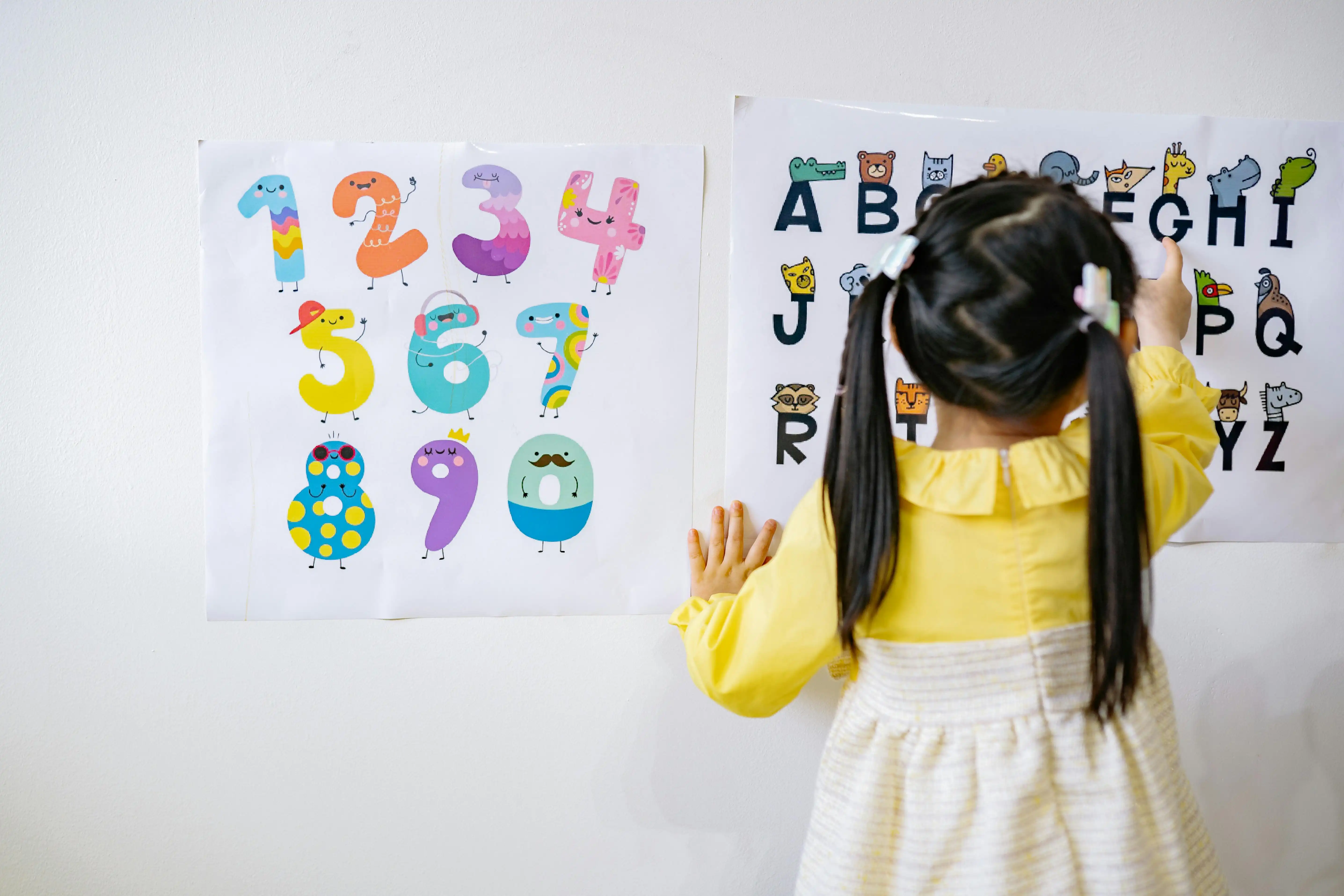 Young child facing a wall with 2 posters on it. One of the alphabet that the child is pointing to and the other has numbers on it.
