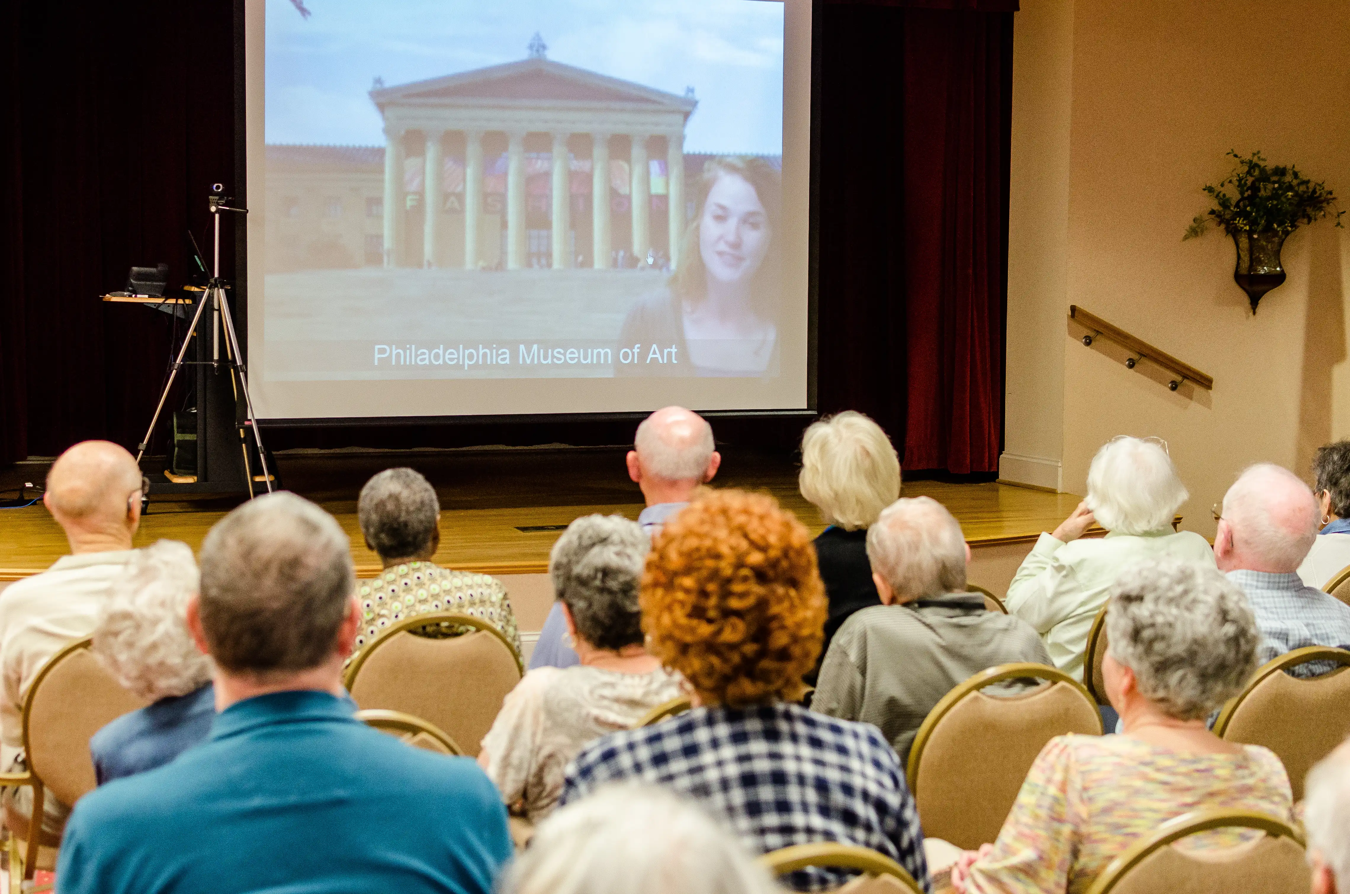 A group of older adults sitting down listening to the presenter on a virtual field trip that is in front of a museum.