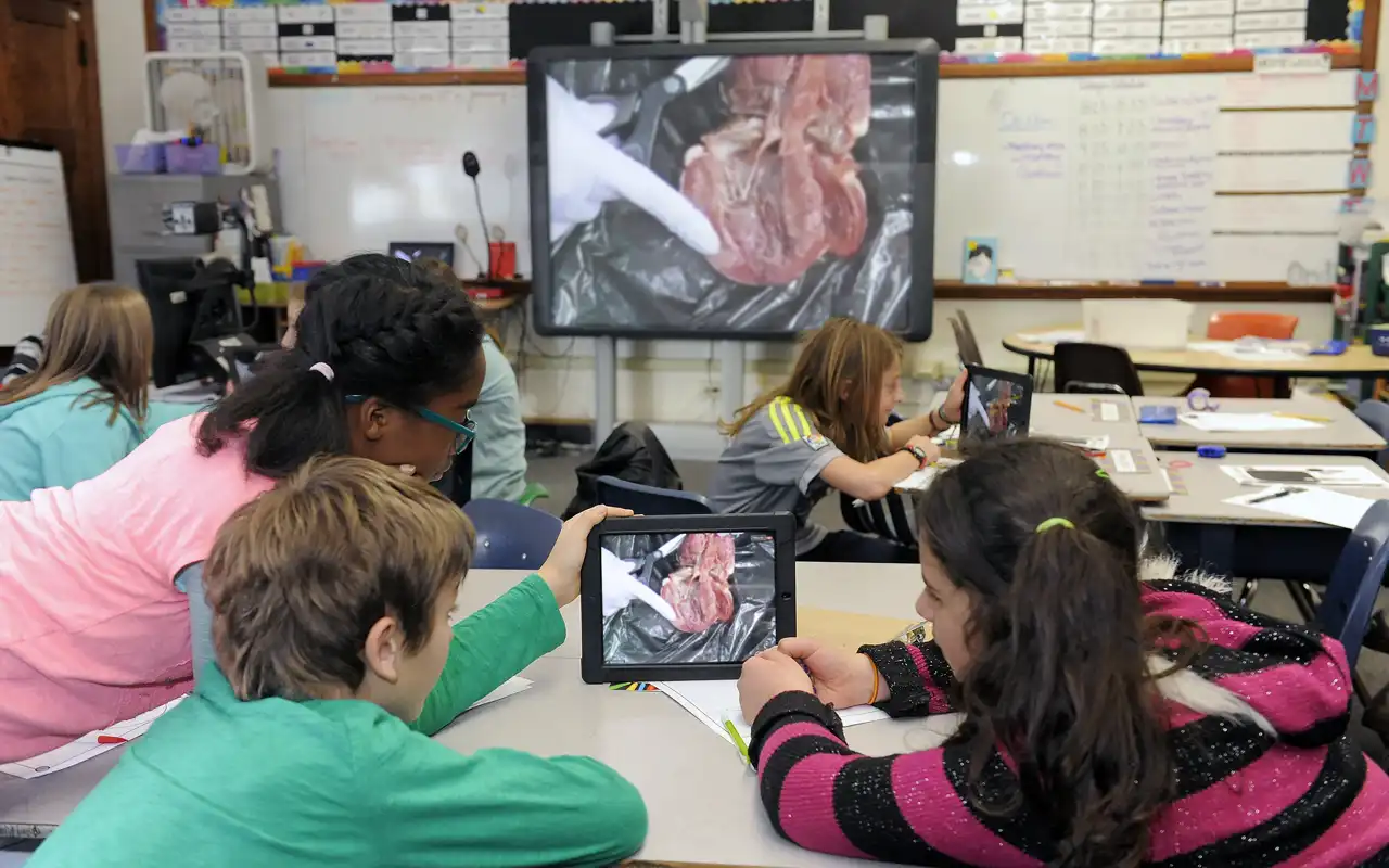 students look at their tablet in the center of a table and at the screen that are both displaying a heart