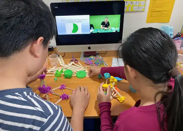 2 students works in a project with blocks and sticks as a presenter is giving instructions on the screen.