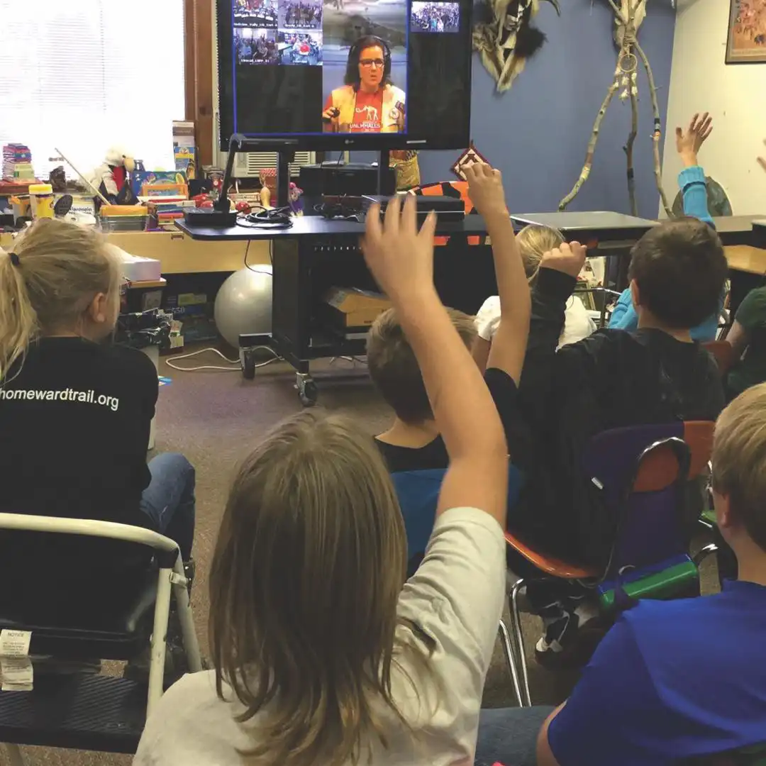 students raising their hands as they get ready to answer a question from the presenter of a virtual field trip on screen