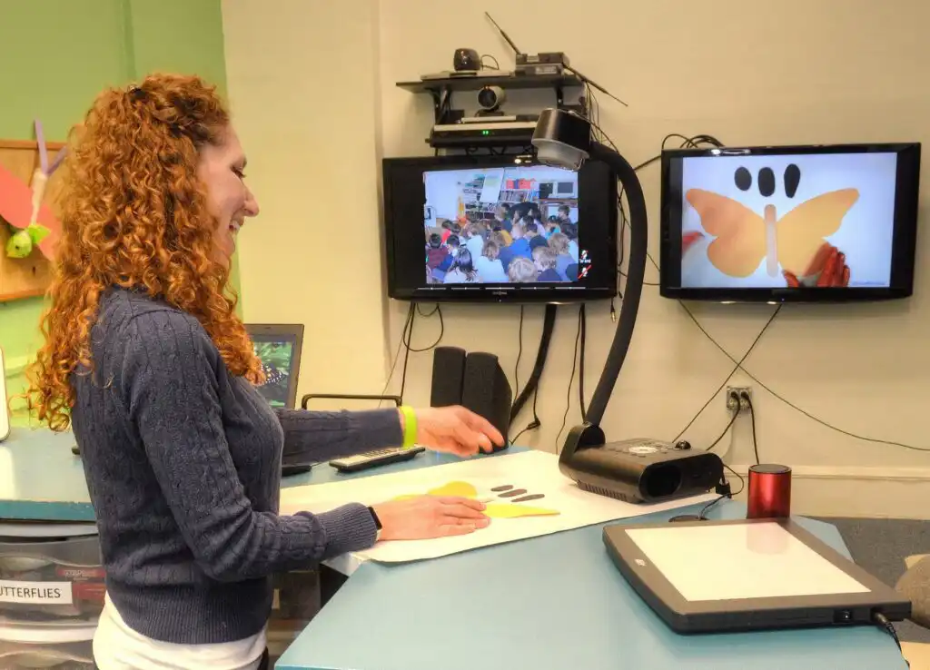 A presenter at the Center for Puppetry arts behind a counter with parts of a butterfly puppet laid out talking to students on one screen with another large screen showing the butterfly parts