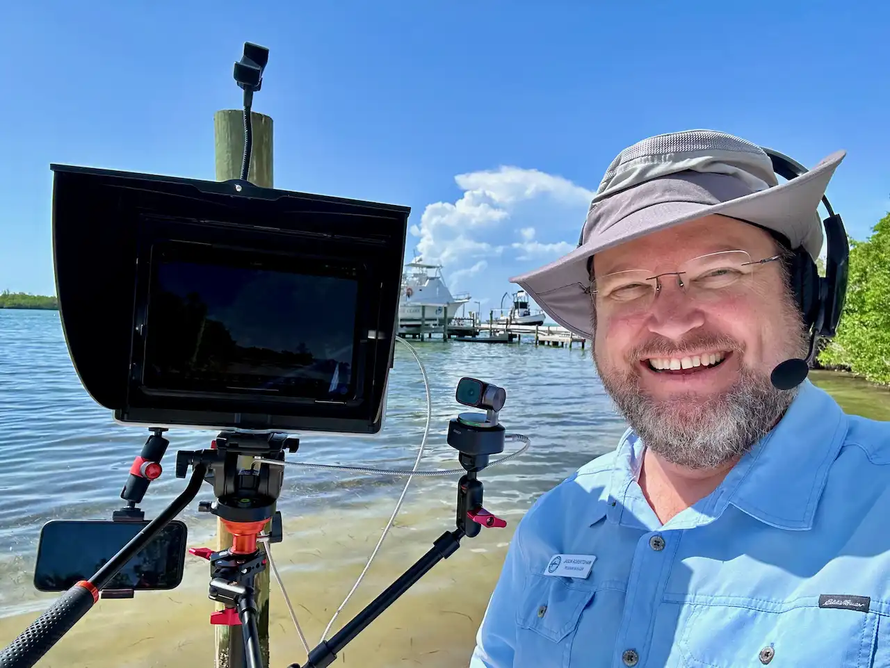 Presenter on the beach with the ocean behind them. The presenter is smiling next to their camera and has a headset on.