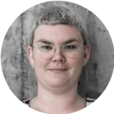Person with short gray hair, glasses, and neutral expression wearing a black top against gray background.