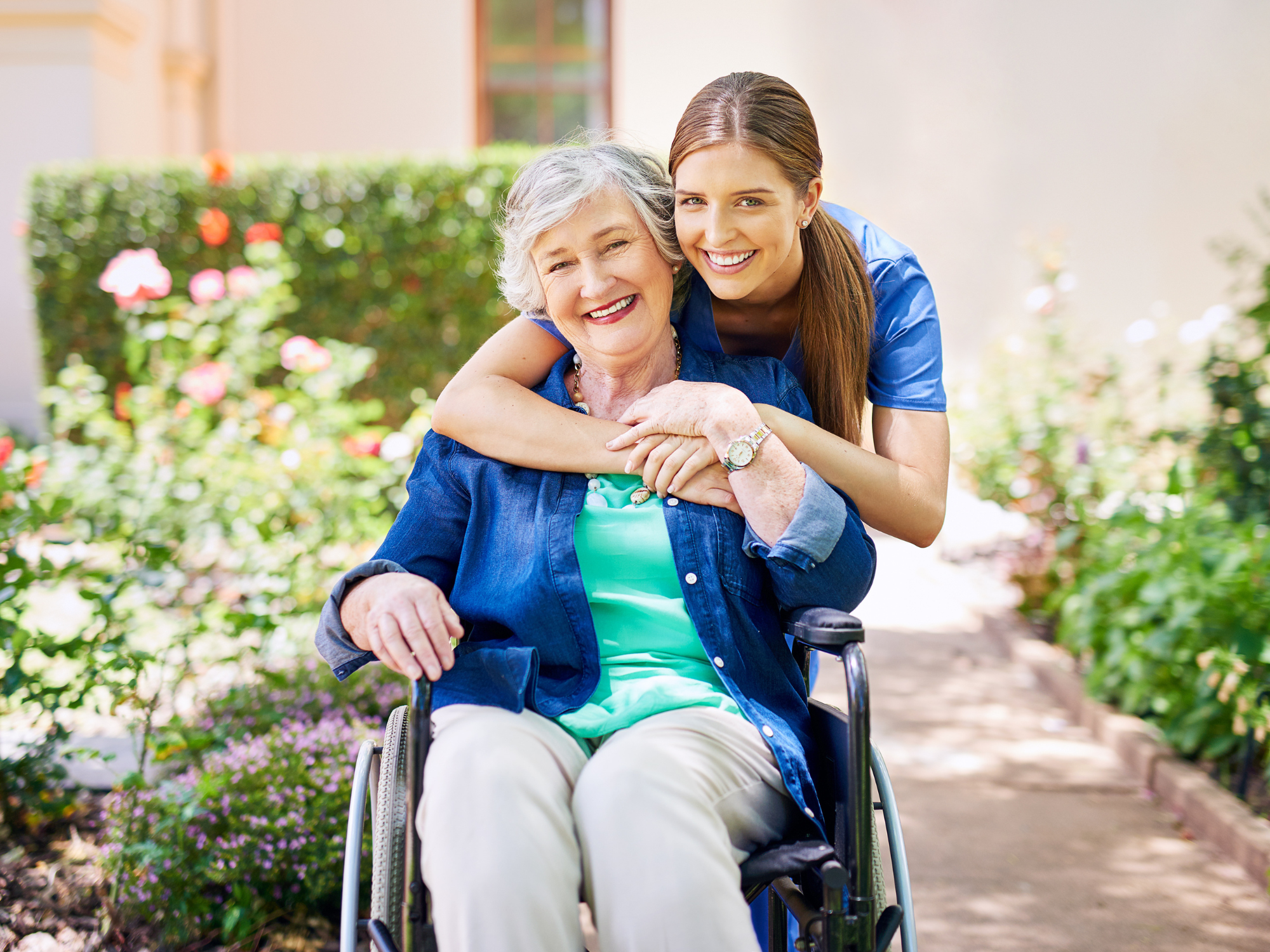 Caregiver in blue scrubs hugging an elderly woman in a wheelchair 