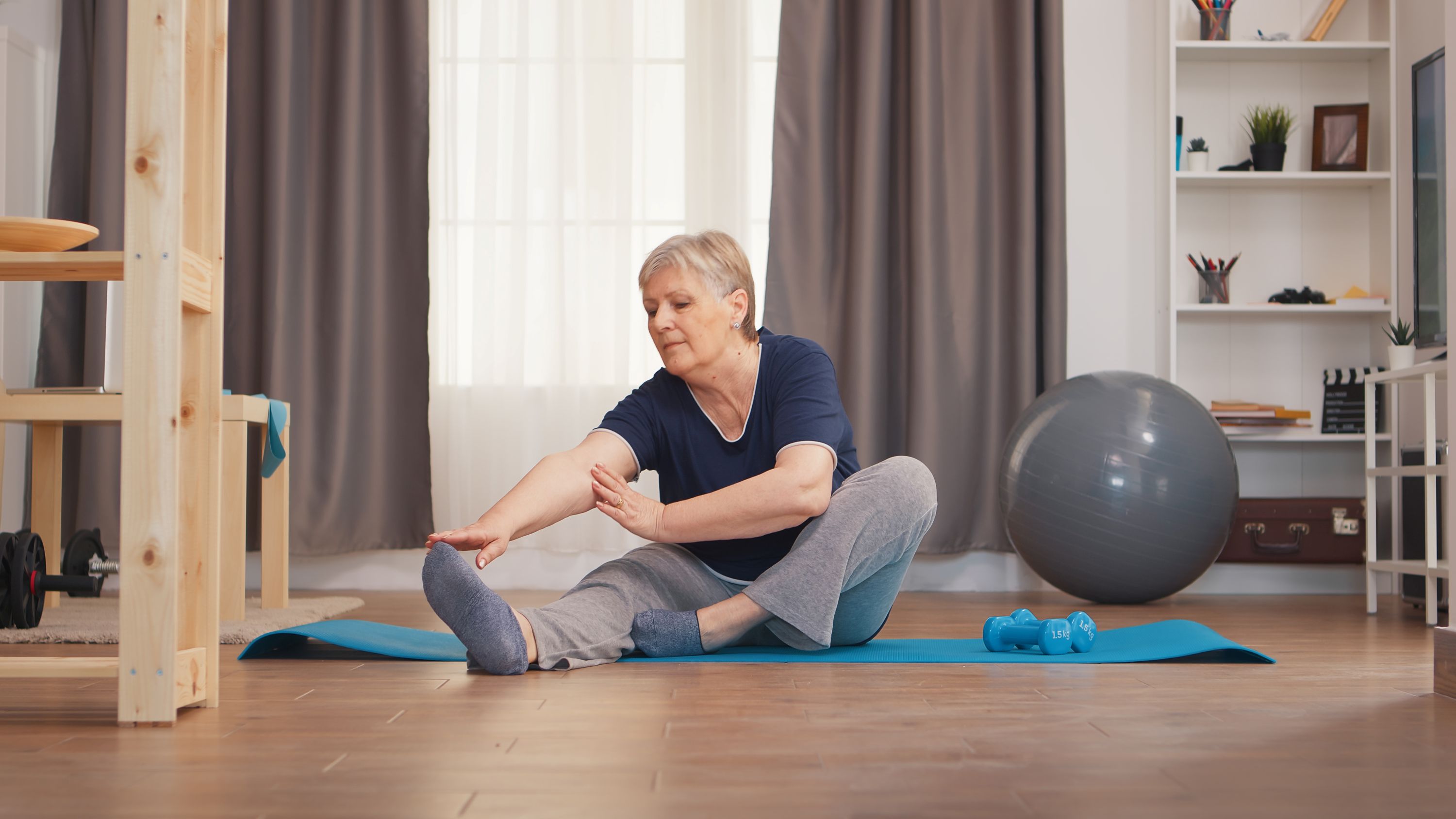Old woman stretching legs sitting on yoga mat in living room. Active healthy lifestyle sporty old person training workout home wellness and indoor exercising