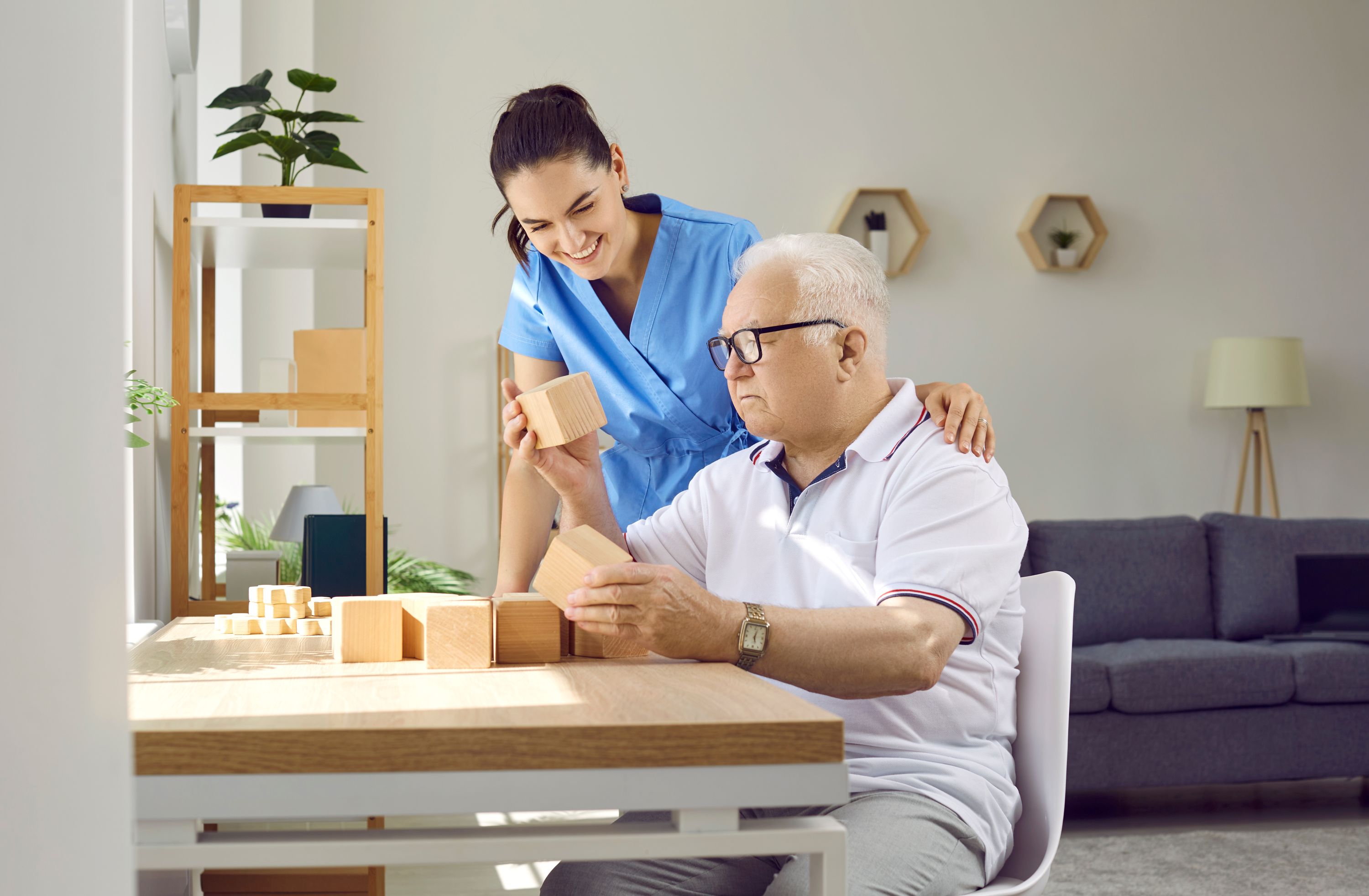 Friendly nurse helping older man in retirement home. Senior man with cognitive problems sitting at desk and building wood block house. Dementia care, people with Alzheimer's disease concept