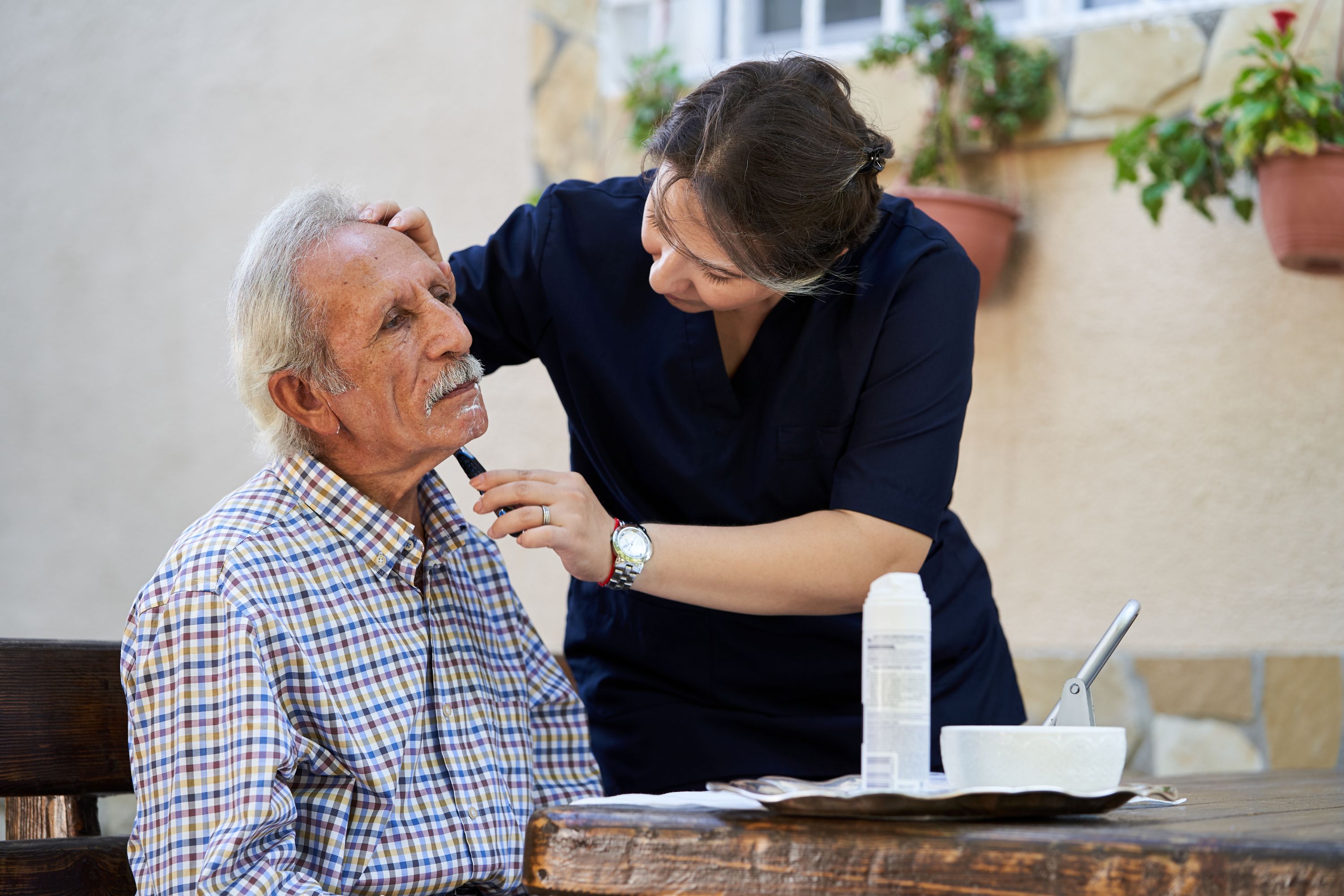 Professional helpful caregiver shaving the face of a senior man during home visit