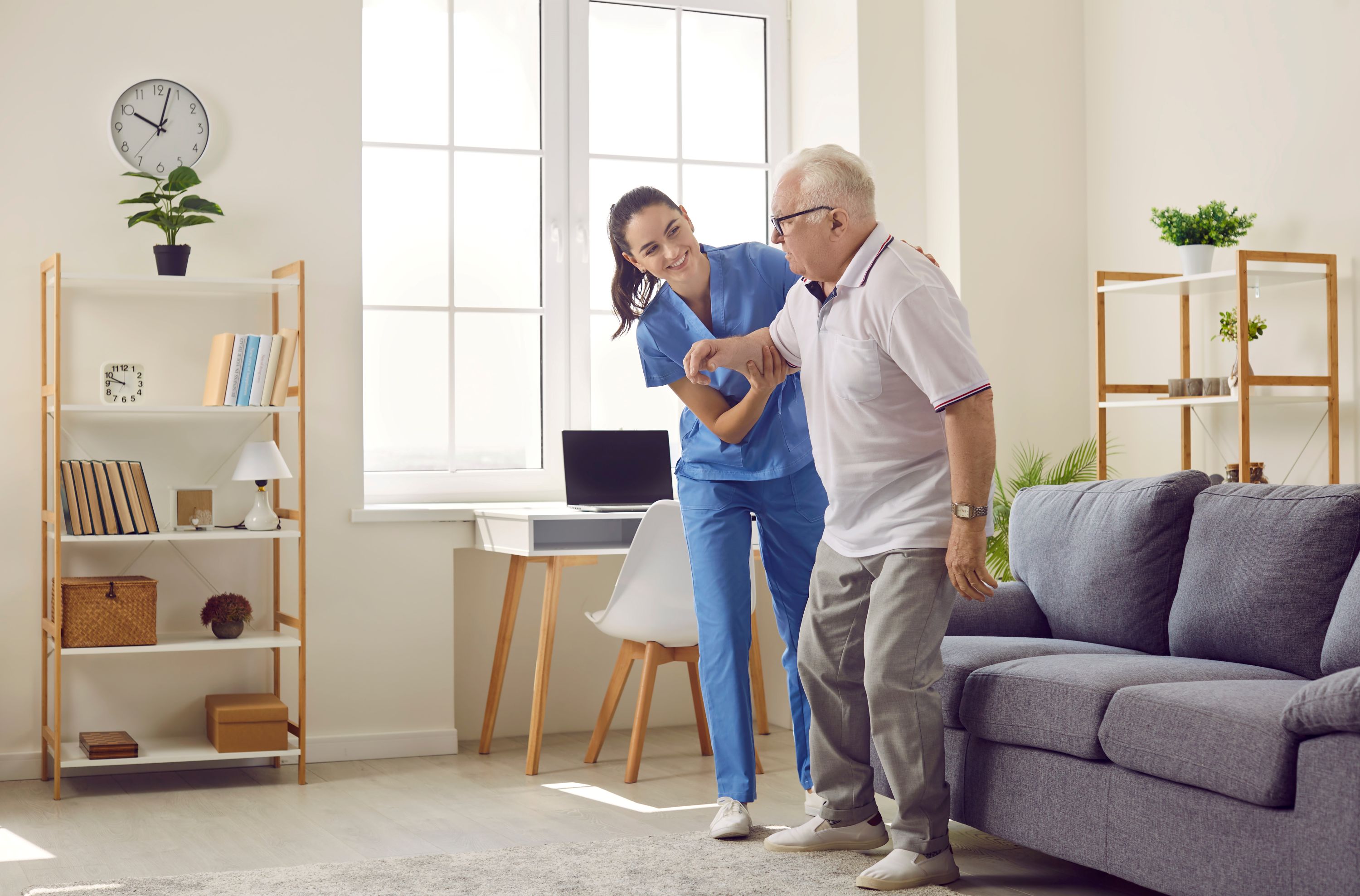 Friendly young female caregiver who cares for elderly man in nursing home helps him get up from sofa.