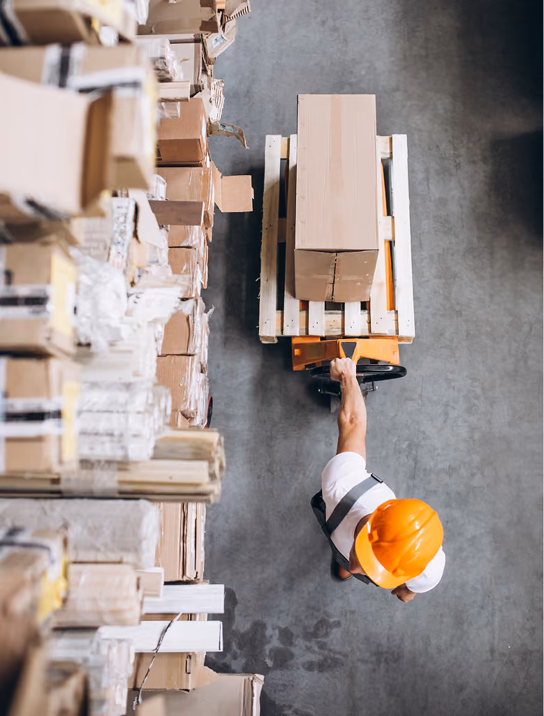 Top view of a worker in an orange helmet moving a pallet jack with a box in a warehouse.