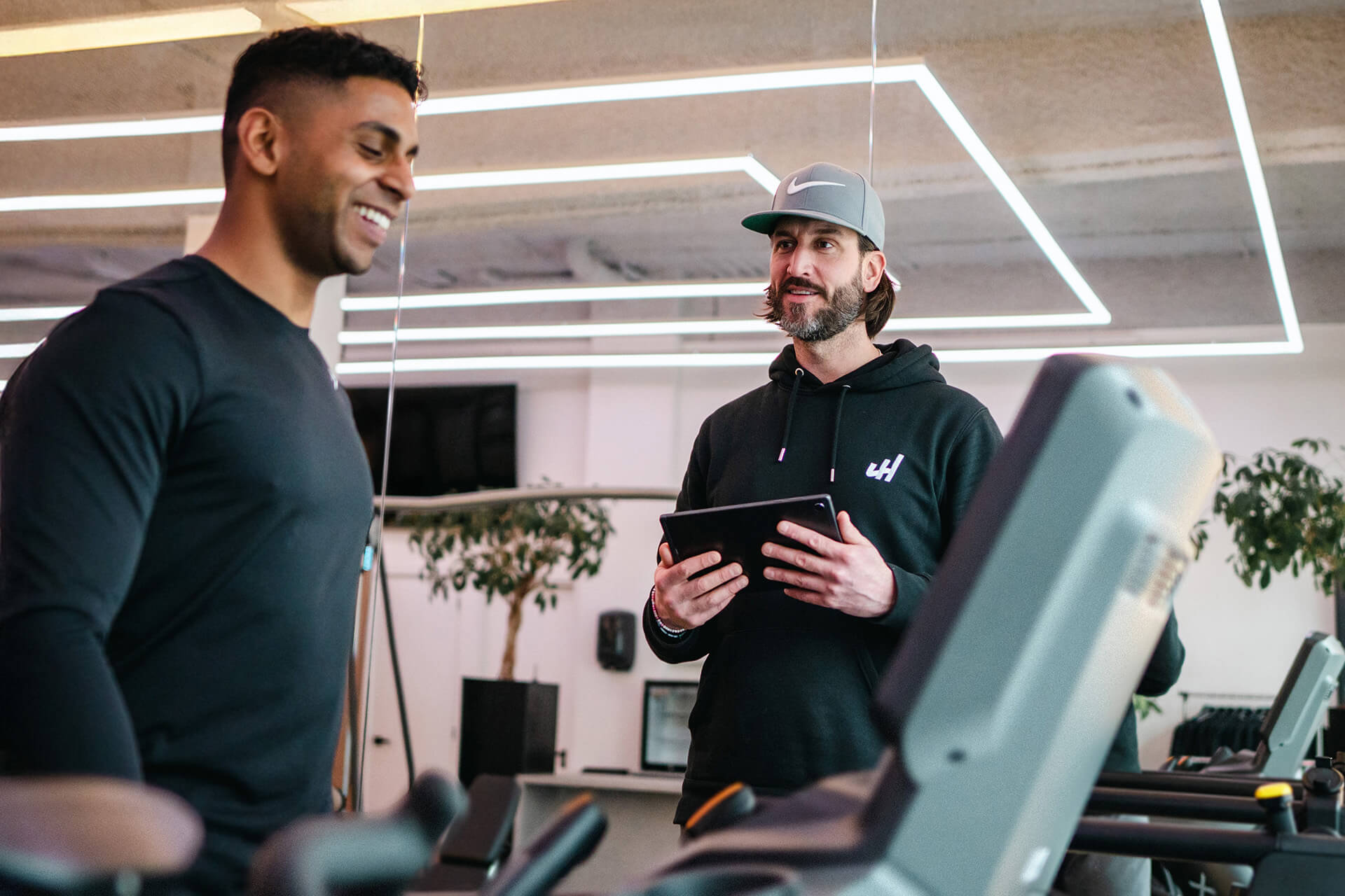 Twee mannen in een sportschool praten; een man met een tablet, de ander lacht terwijl hij op een loopband staat.