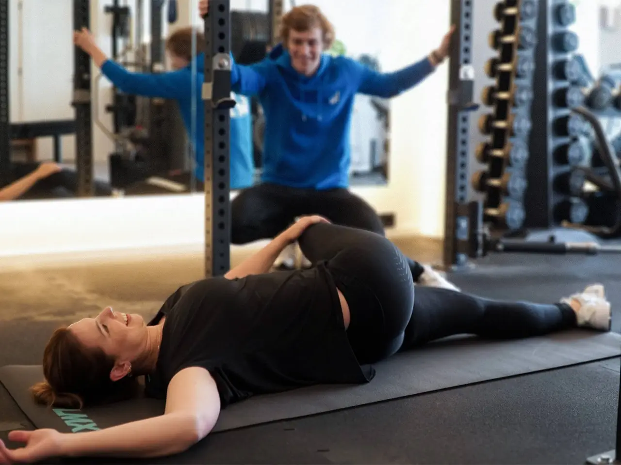 Vrouw die op een mat ligt en een rugstretch doet terwijl een man haar begeleidt in een sportschool.