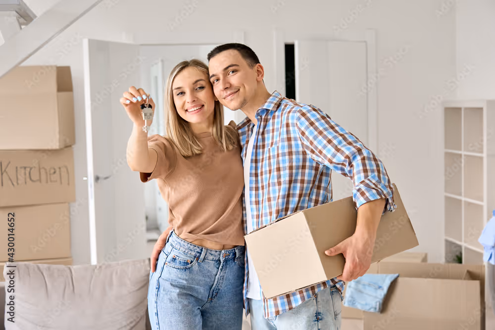 A couple smiling, holding keys and a box, standing in their new home.