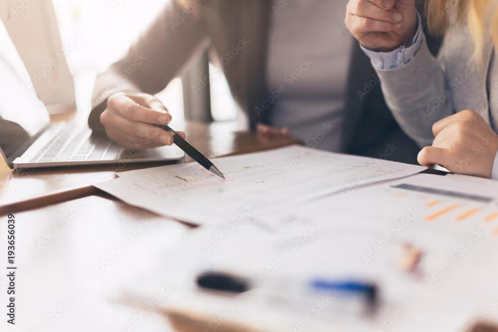Two people review documents at a desk, one pointing with a pen towards printed charts.