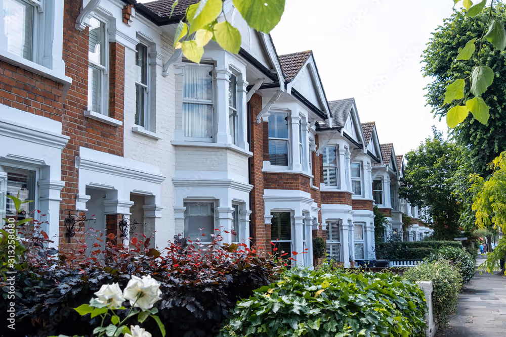 Row of similar brick houses with white trim, framed by bushes and trees along the sidewalk.