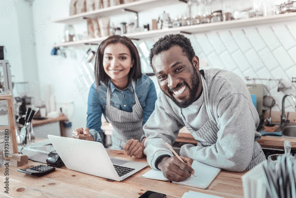 Two people in aprons smiling at a desk with a laptop and notepad.