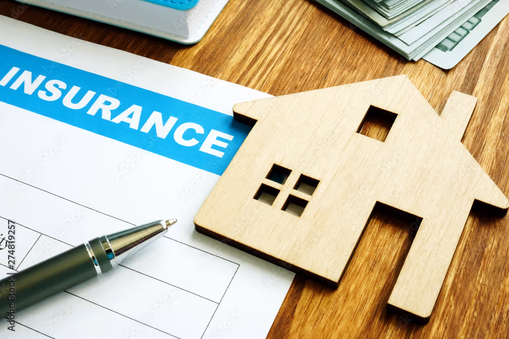 A wooden house model sits on an insurance form next to a pen on a table.