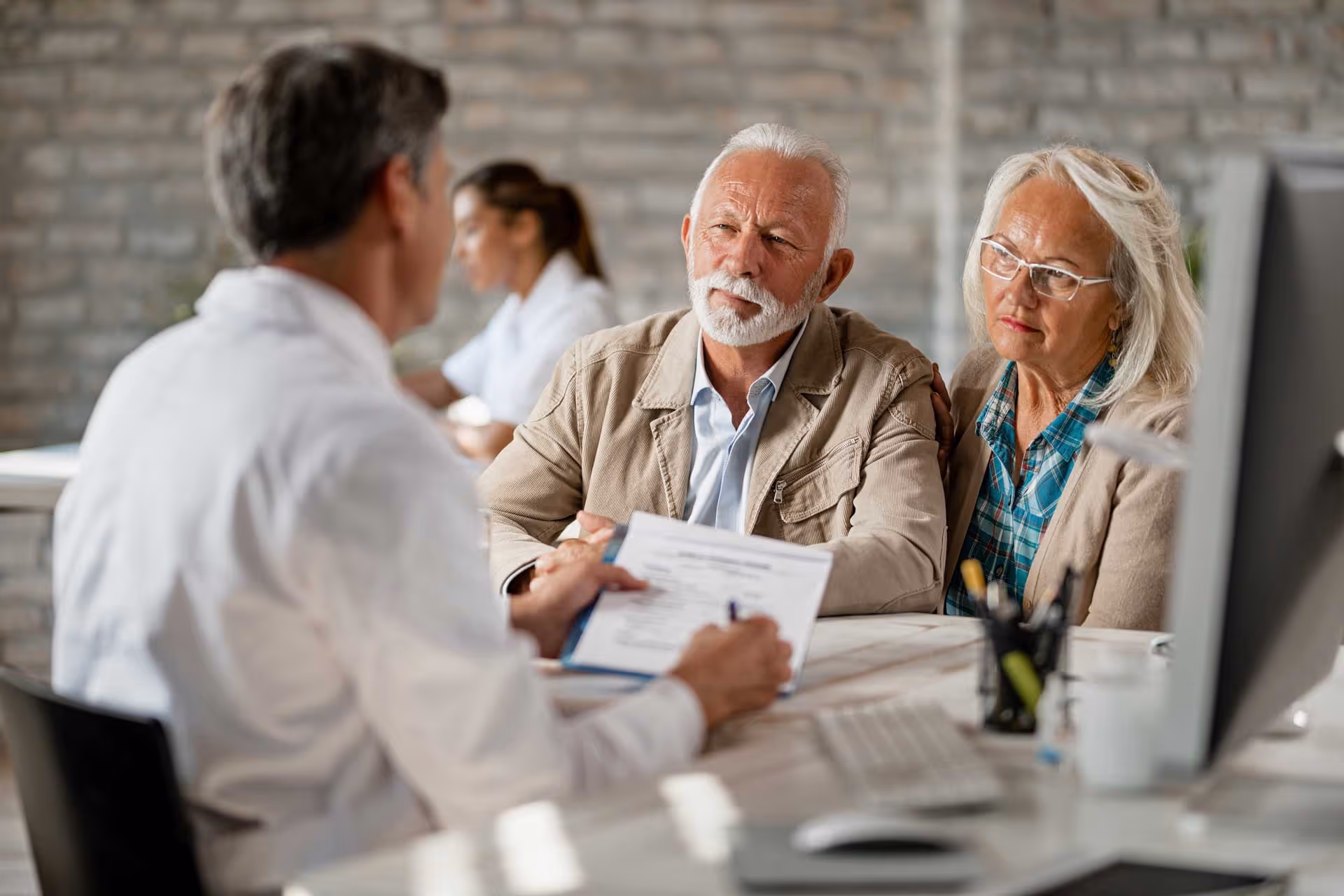 A doctor discusses a medical document with an elderly couple sitting across a desk in an office.