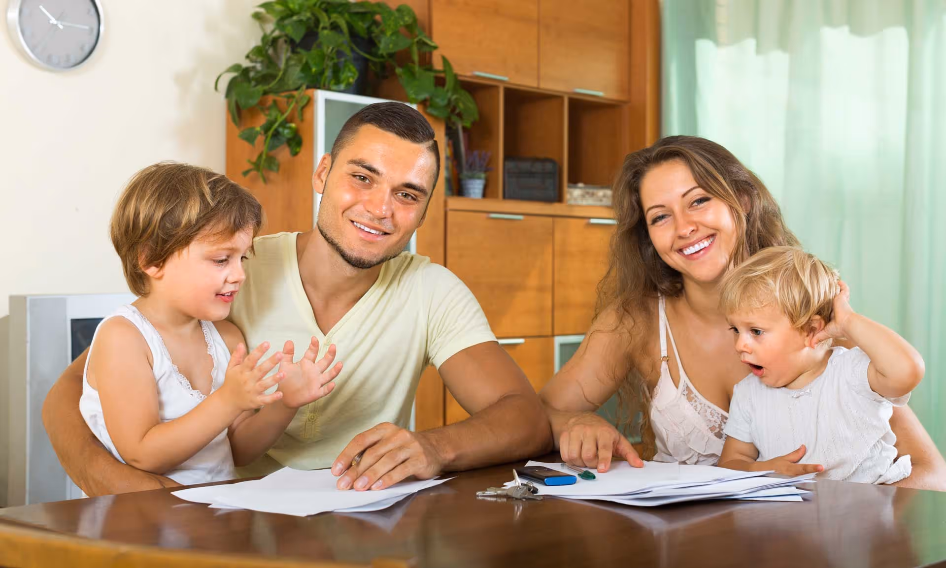 A smiling family sits at a table with papers and a pen, engaging with two small children.