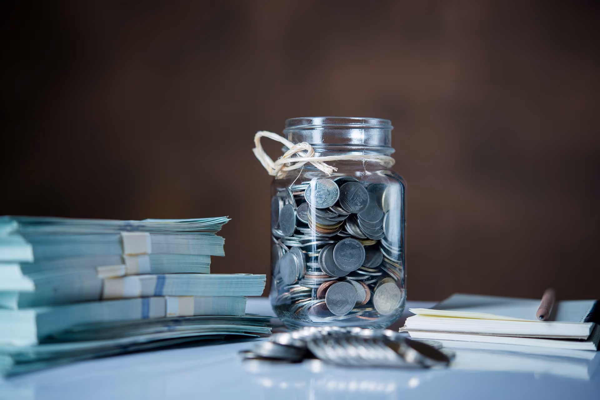 A jar filled with coins on a table beside stacked bundles of cash and a notebook.