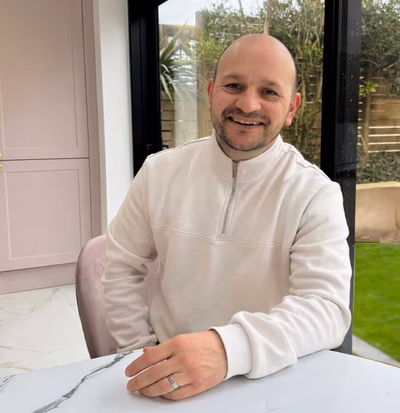 Michael sitting at a table, smiling, wearing a white sweater with a ring on his finger.