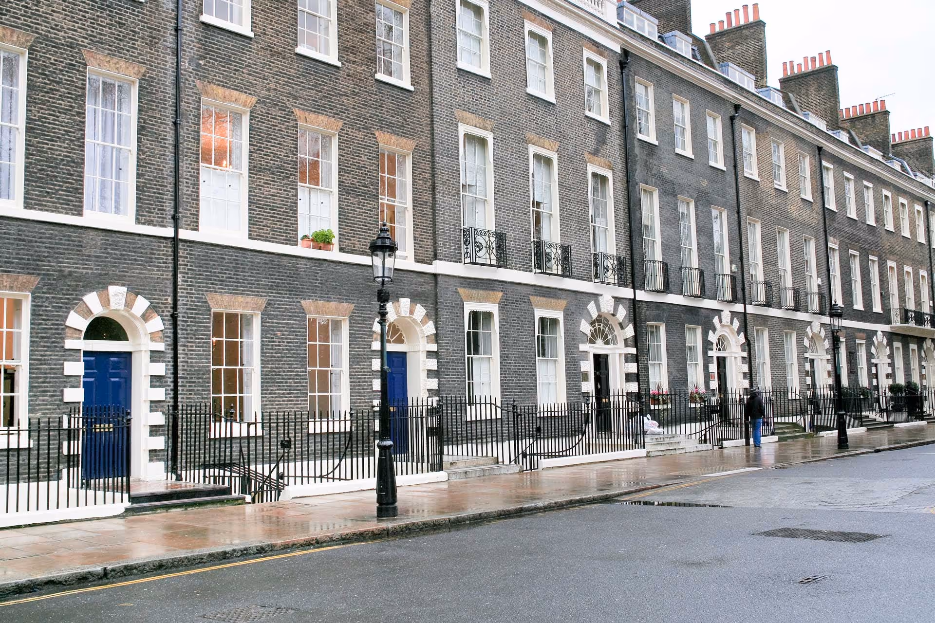 A row of classic brick townhouses with arched windows and blue doors on a wet street.