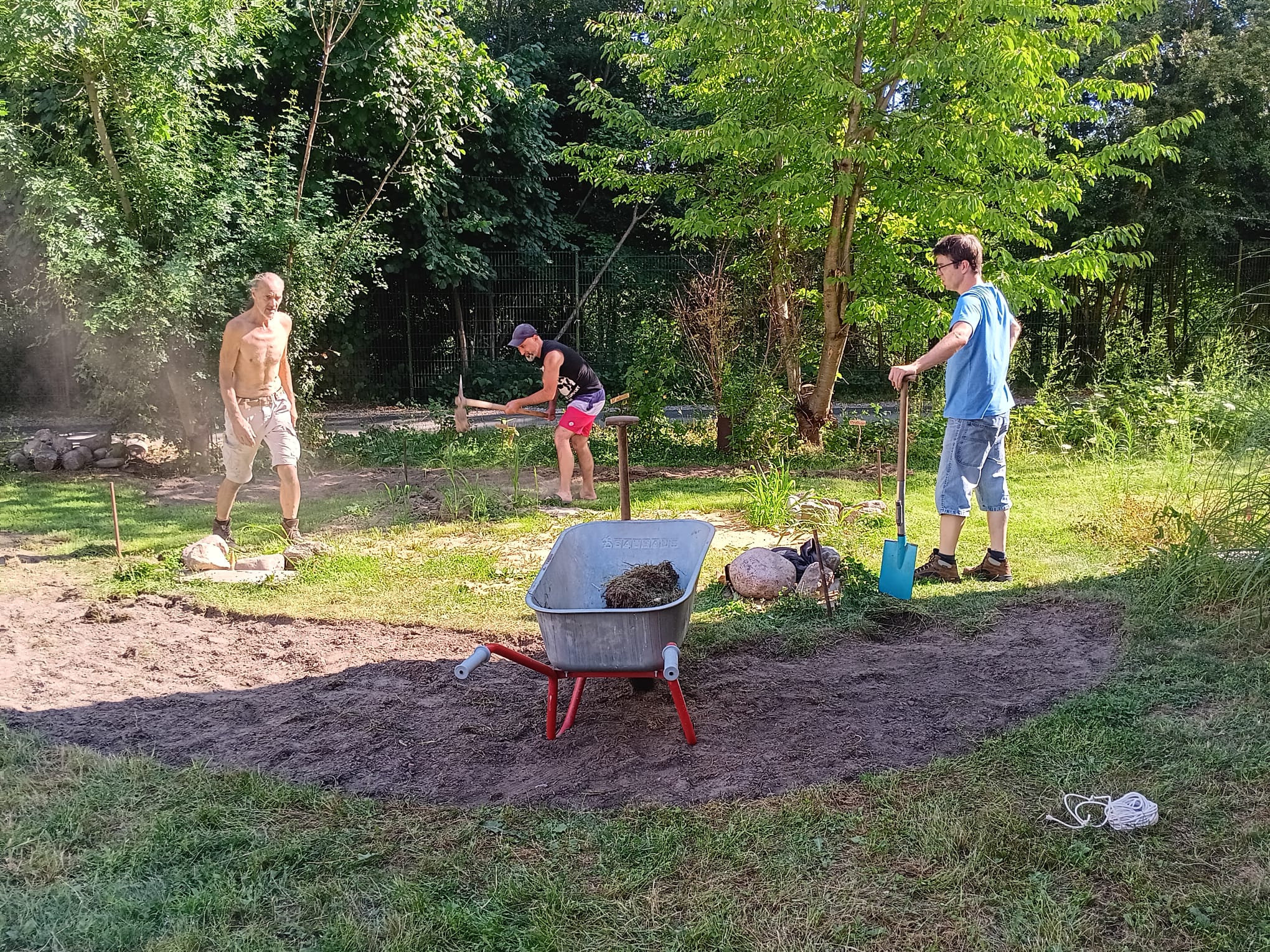 Mehrere Menschen bereiten gemeinsam eine künftige Spielfläche im Grünen vor; zu sehen sind Schubkarre, Spaten und die abgesteckte Bodenfläche.