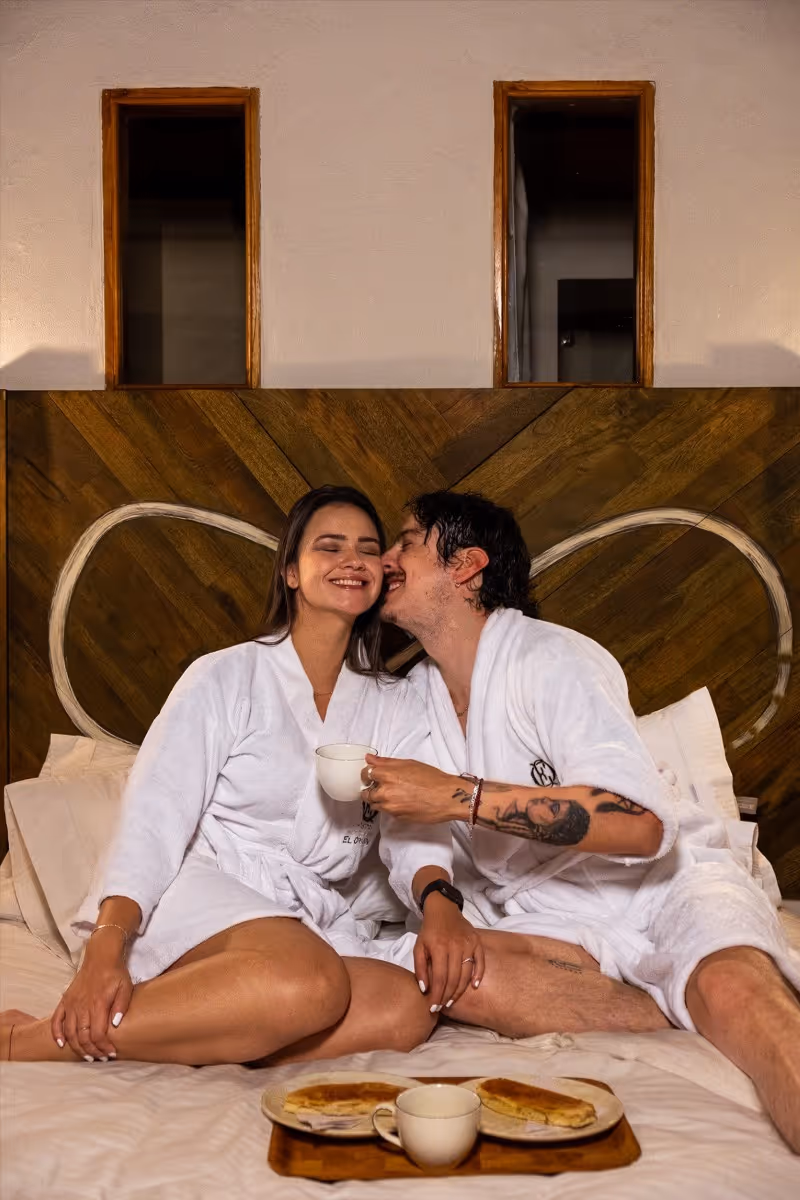 Pareja disfrutando una escapada romántica en una habitación del Hotel 1572 El Origen en Villa de Leyva, con desayuno en la cama y ambiente cálido de descanso y desconexión.