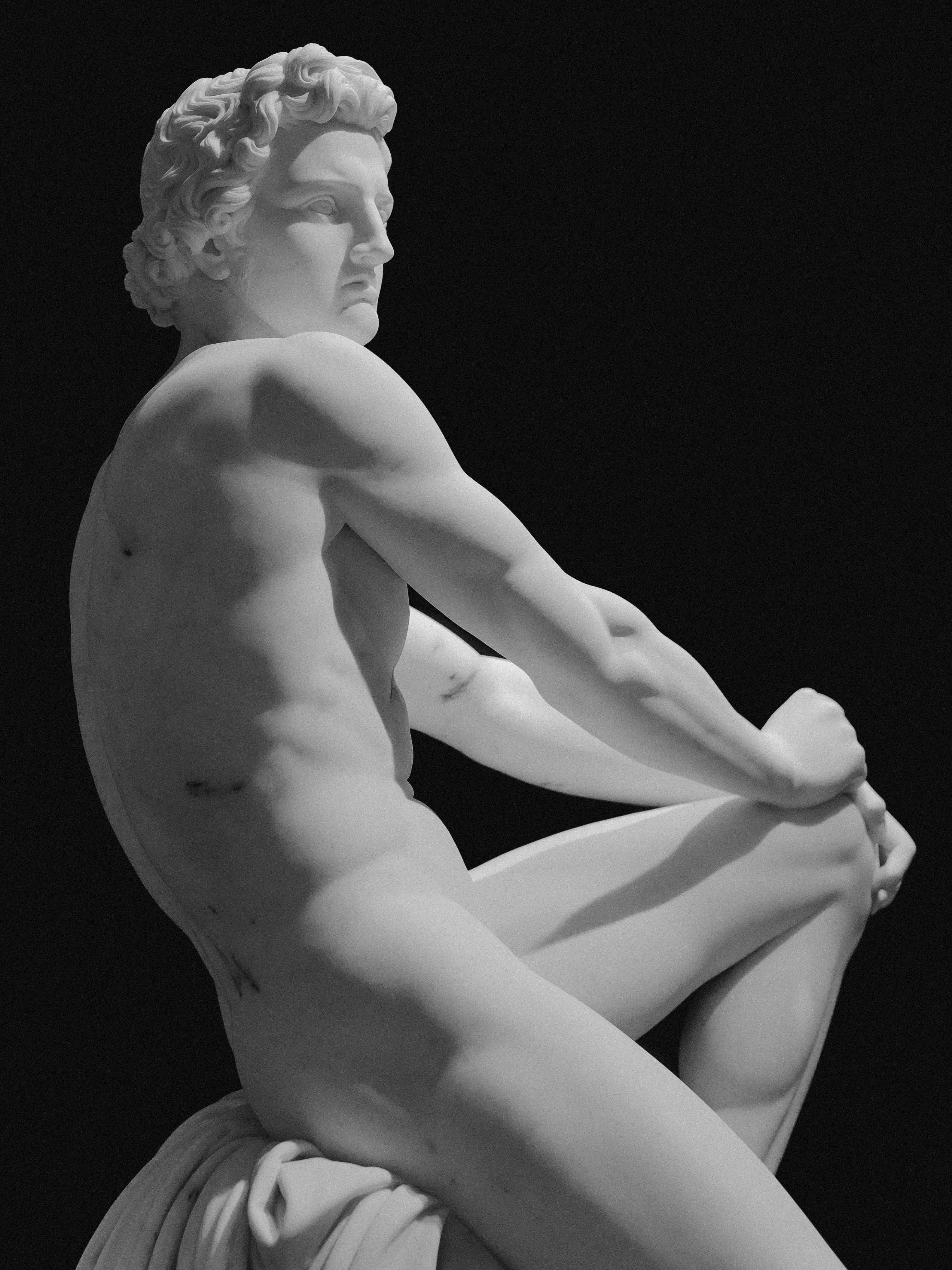 Side view of a classical roman white marble statue of a seated muscular male figure against a black background.
