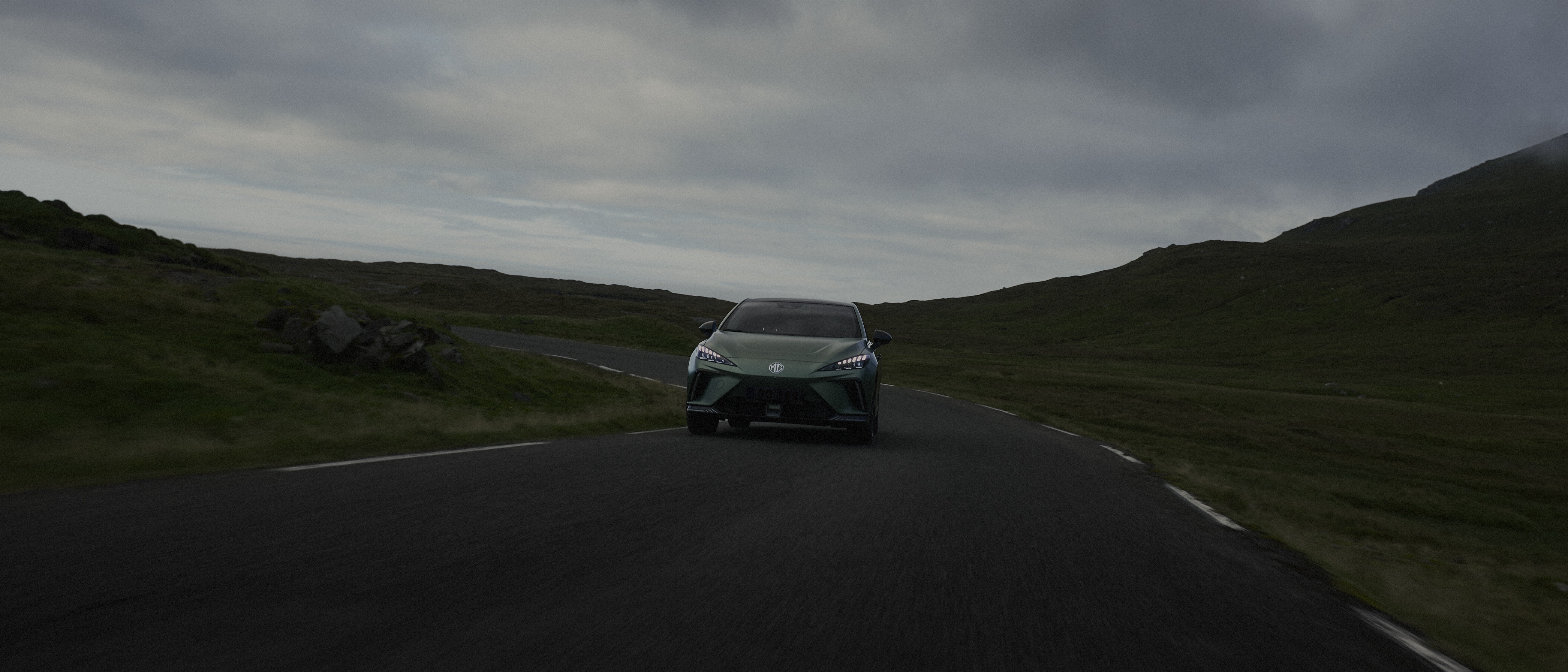 Green MG car driving on a winding road surrounded by grassy hills under a cloudy sky on the Faroe Islands.