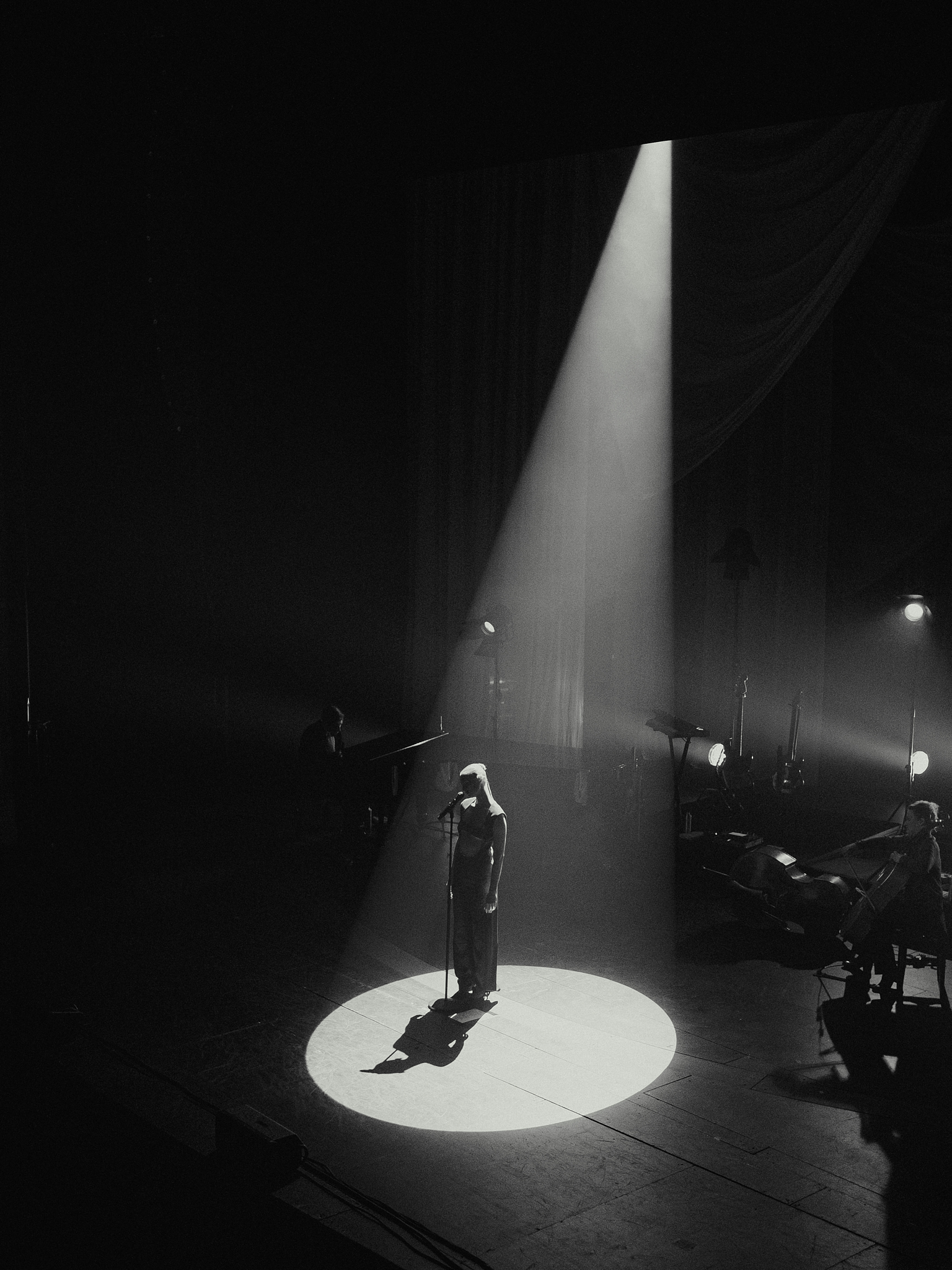 A singer standing alone in a spotlight on stage with musicians playing a piano and cello in dim light.