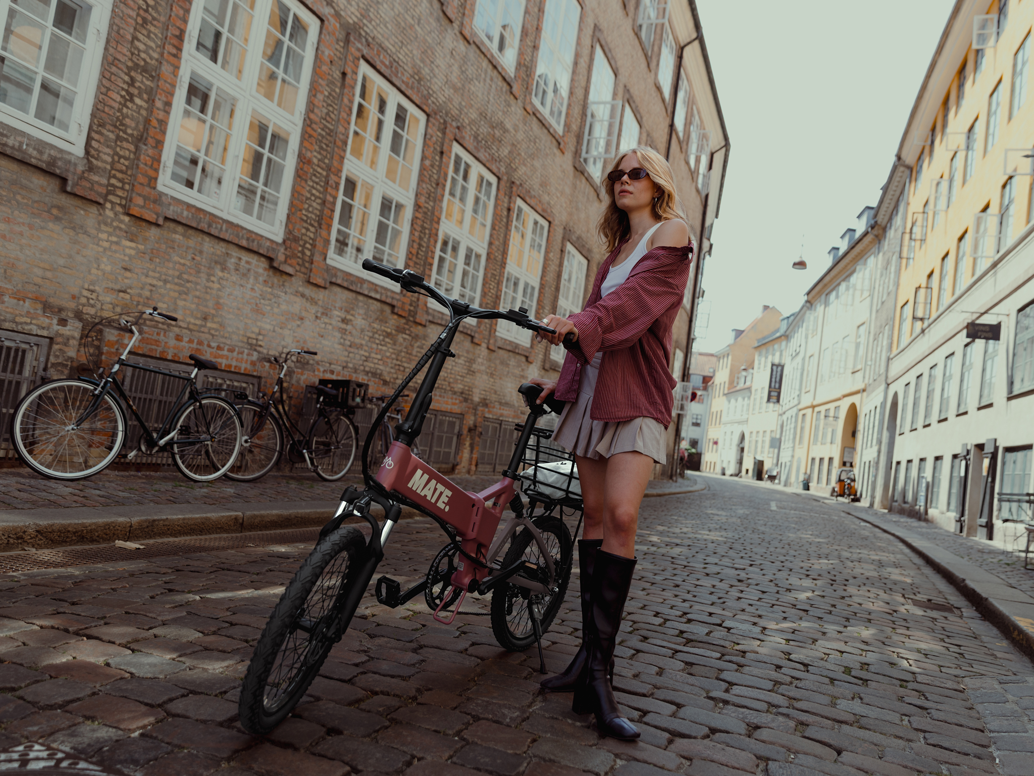 Woman wearing sunglasses and boots standing on a cobblestone street holding a pink MATE electric bike.