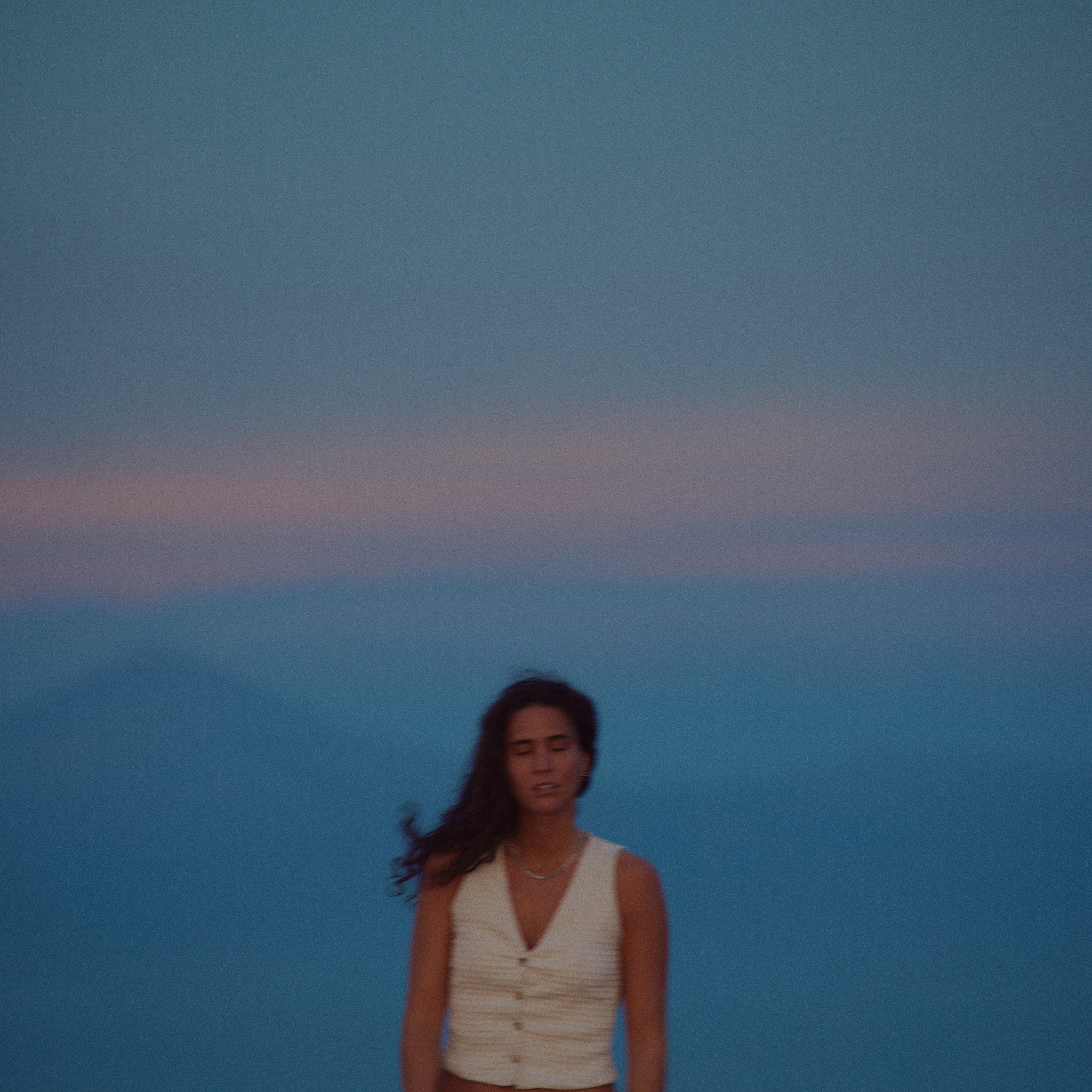 Woman with long dark hair and white cream buttoned vest standing with closed eyes against a blue mountainous background at dusk.