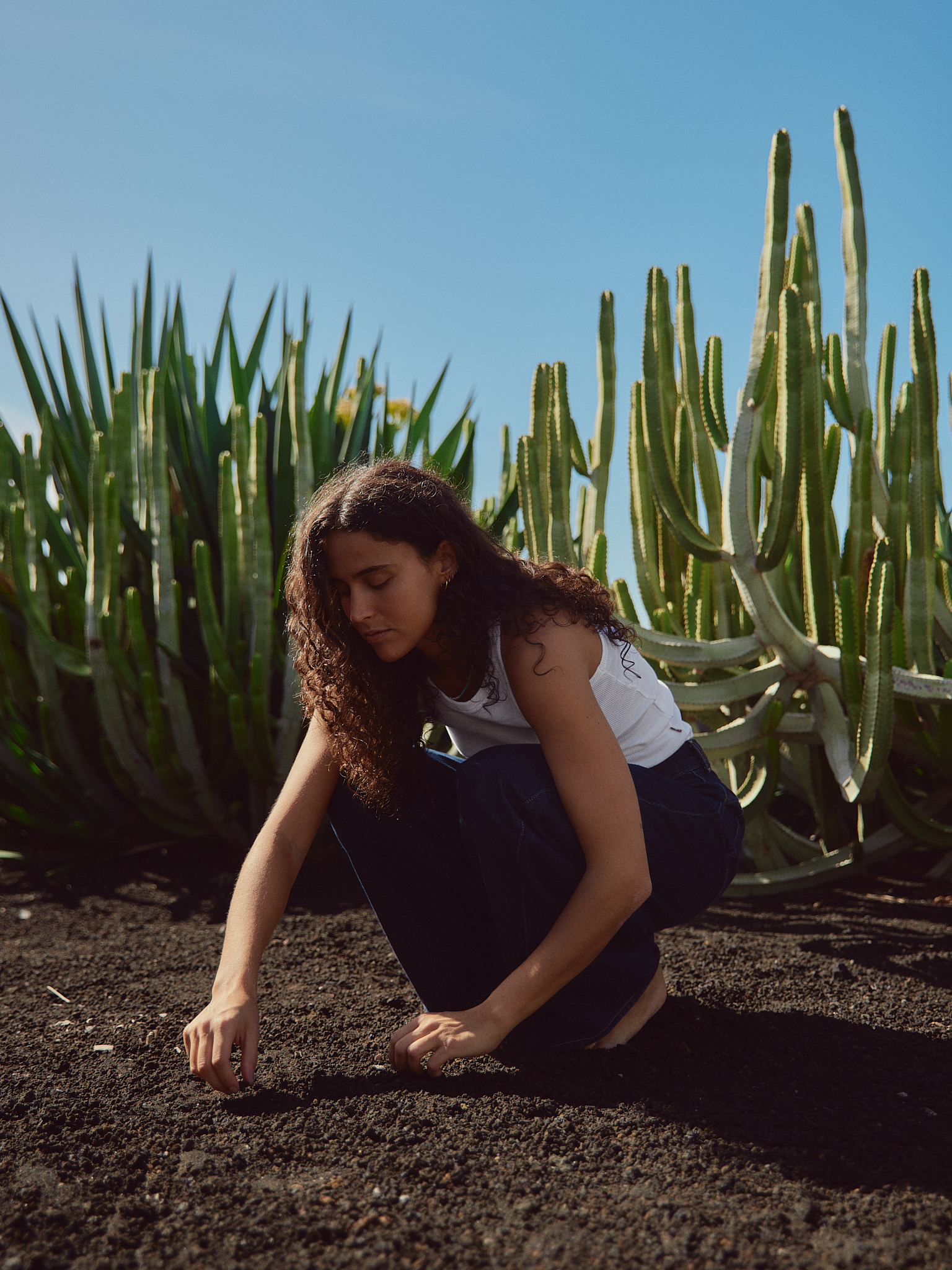 Woman with curly hair crouching on soil in front of tall cacti under clear blue sky.