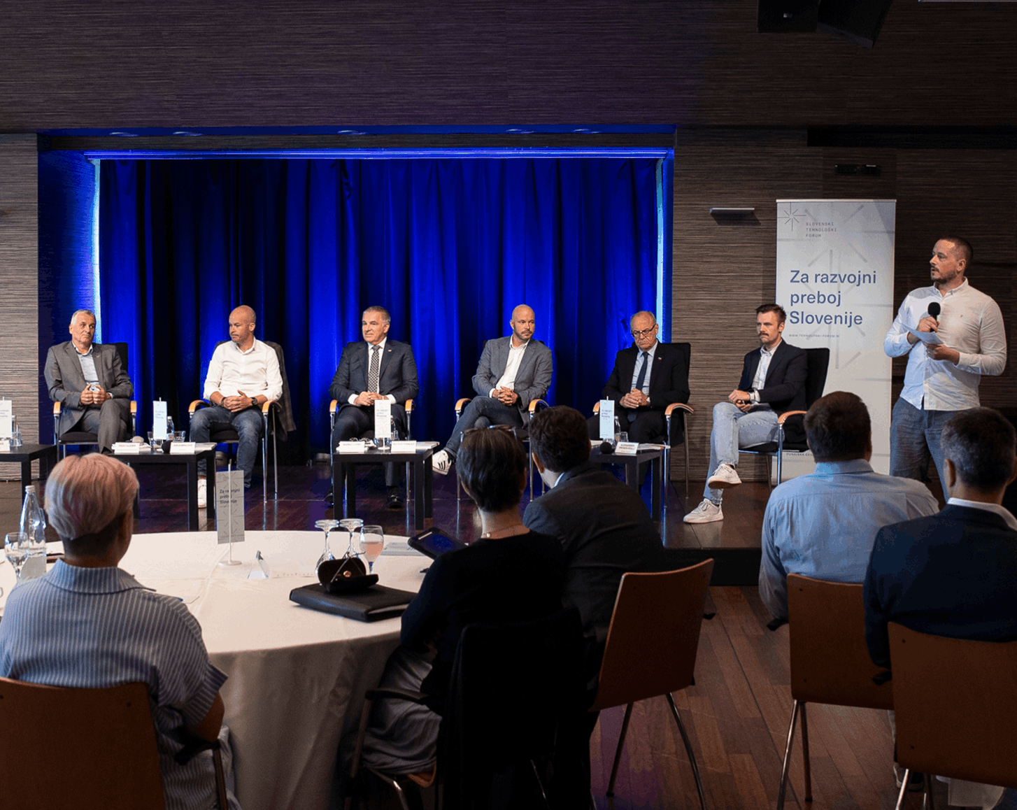Diskusija s šestimi moškimi na odru pred moder publiko, eden stoji in govori v mikrofon, ozadje modra zavesa in napis Za razvojni preboj Slovenije.