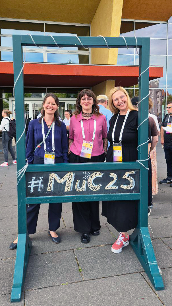 Stefanie Dreiack, Miriam Grünz und Claudia Loitsch stehen nebeneinander hinter einem großen, aufgestellten, blaugrauen Rahmen, auf den mit Kreide groß "#MuC25" geschrieben wurde. Die drei Frauen blicken lächelnd durch den Rahmen zur Kamera.