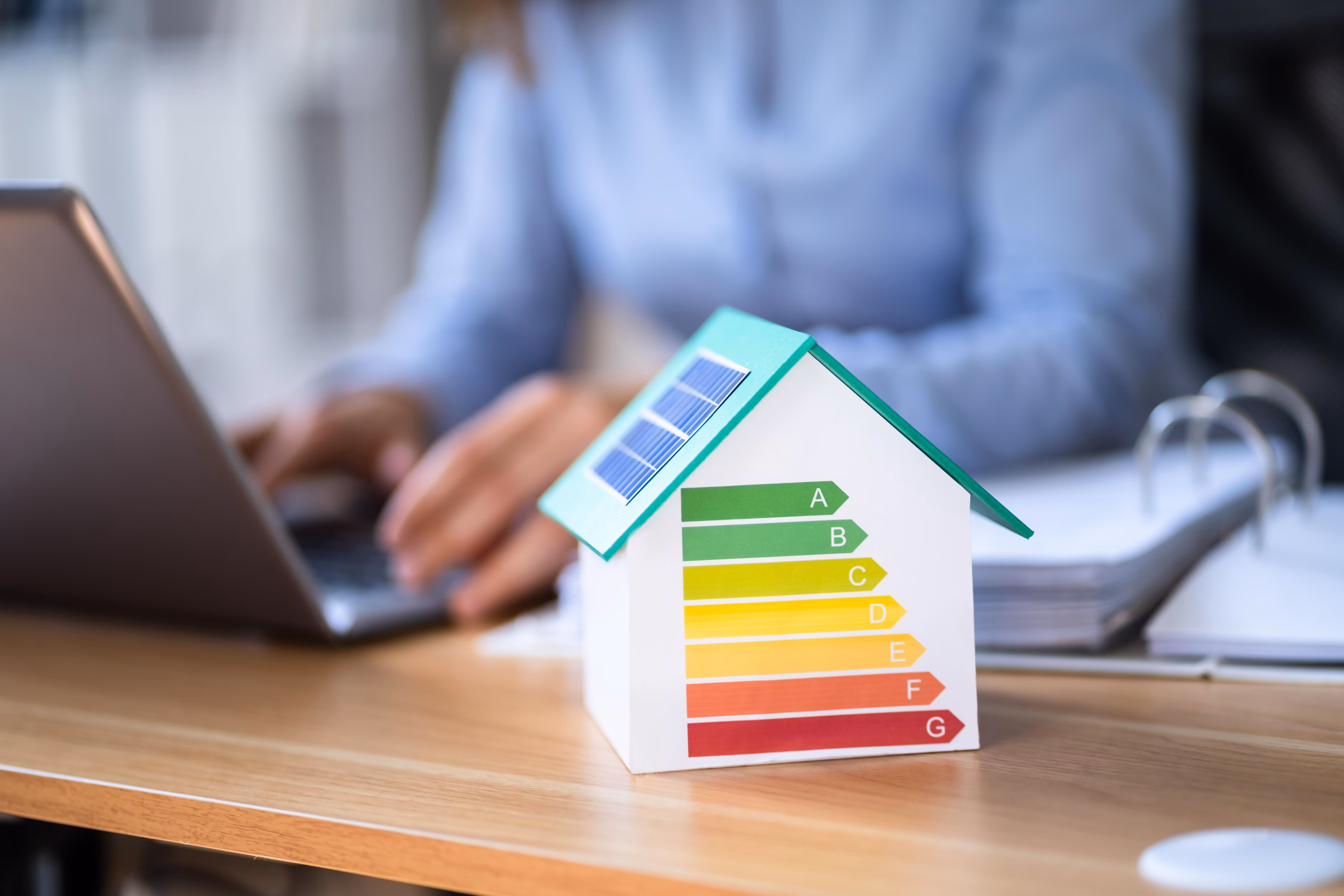 A person works on a laptop next to a small model house with energy efficiency ratings.