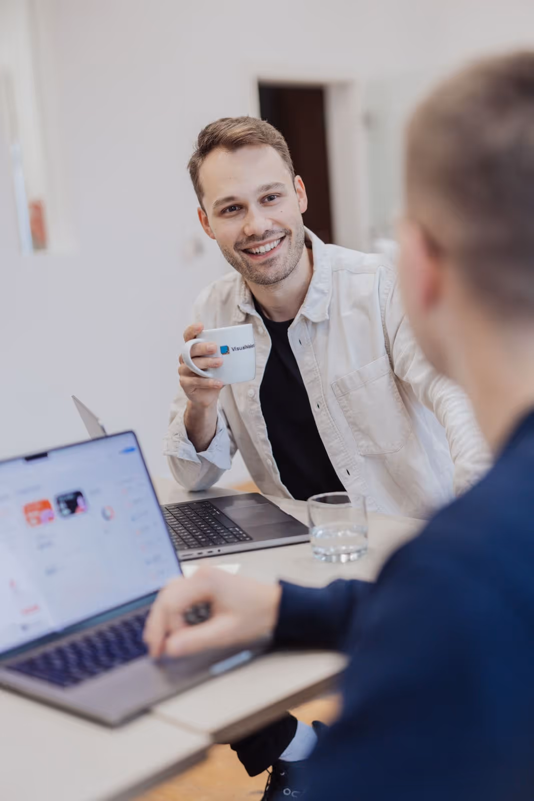 two people in conversation sitting at a table with laptops, one person smiling holding a cup