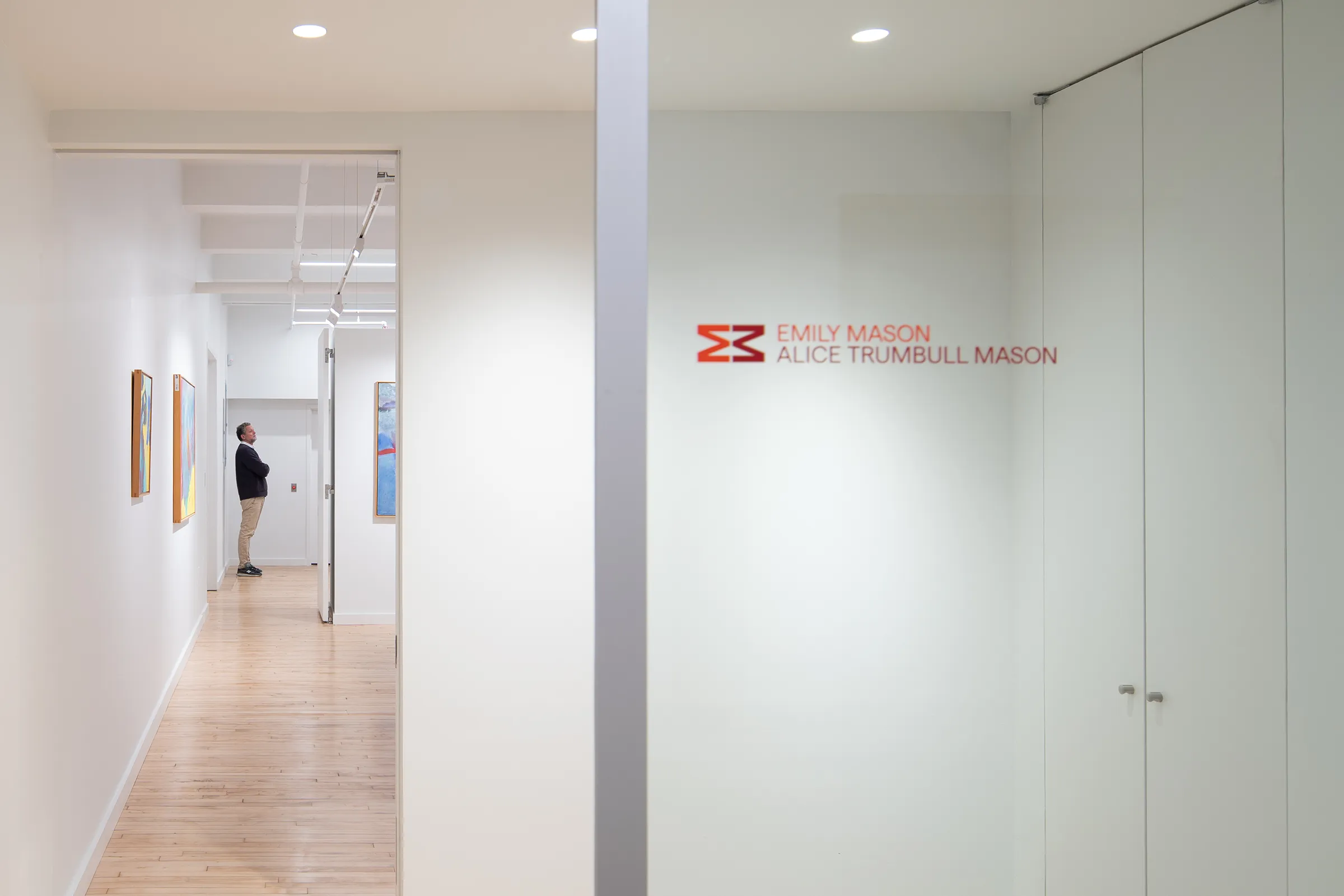modern office, which doubles as a gallery space for EMATM. In the background a man looks at a painting on a wall, in the foreground, a large glass window shows off the EMATM logo
