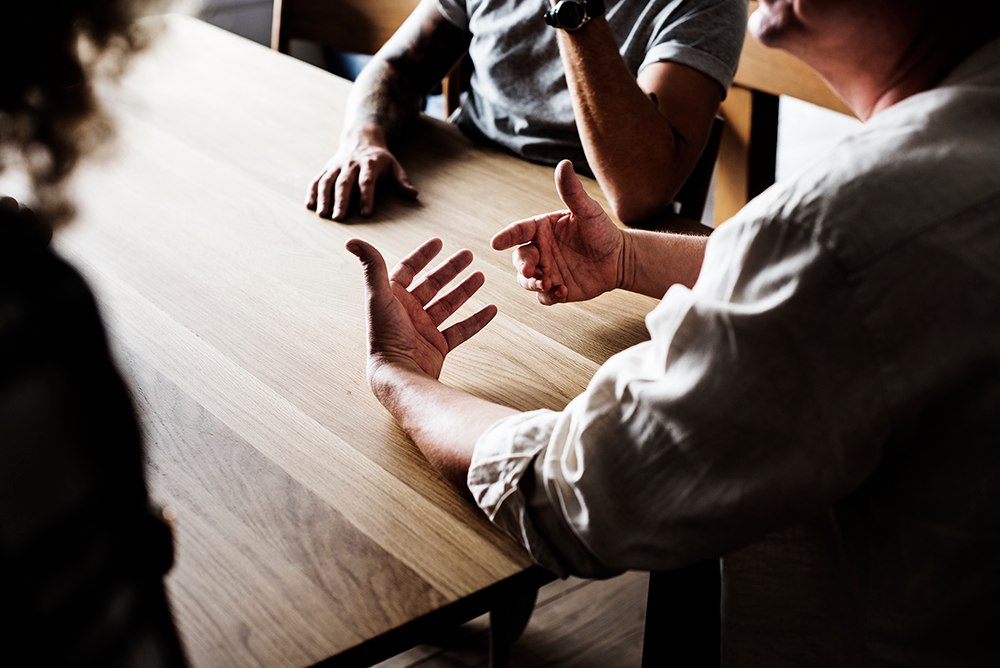 Close-up of three people sitting at a wooden table, one gesturing with open hands during a discussion.