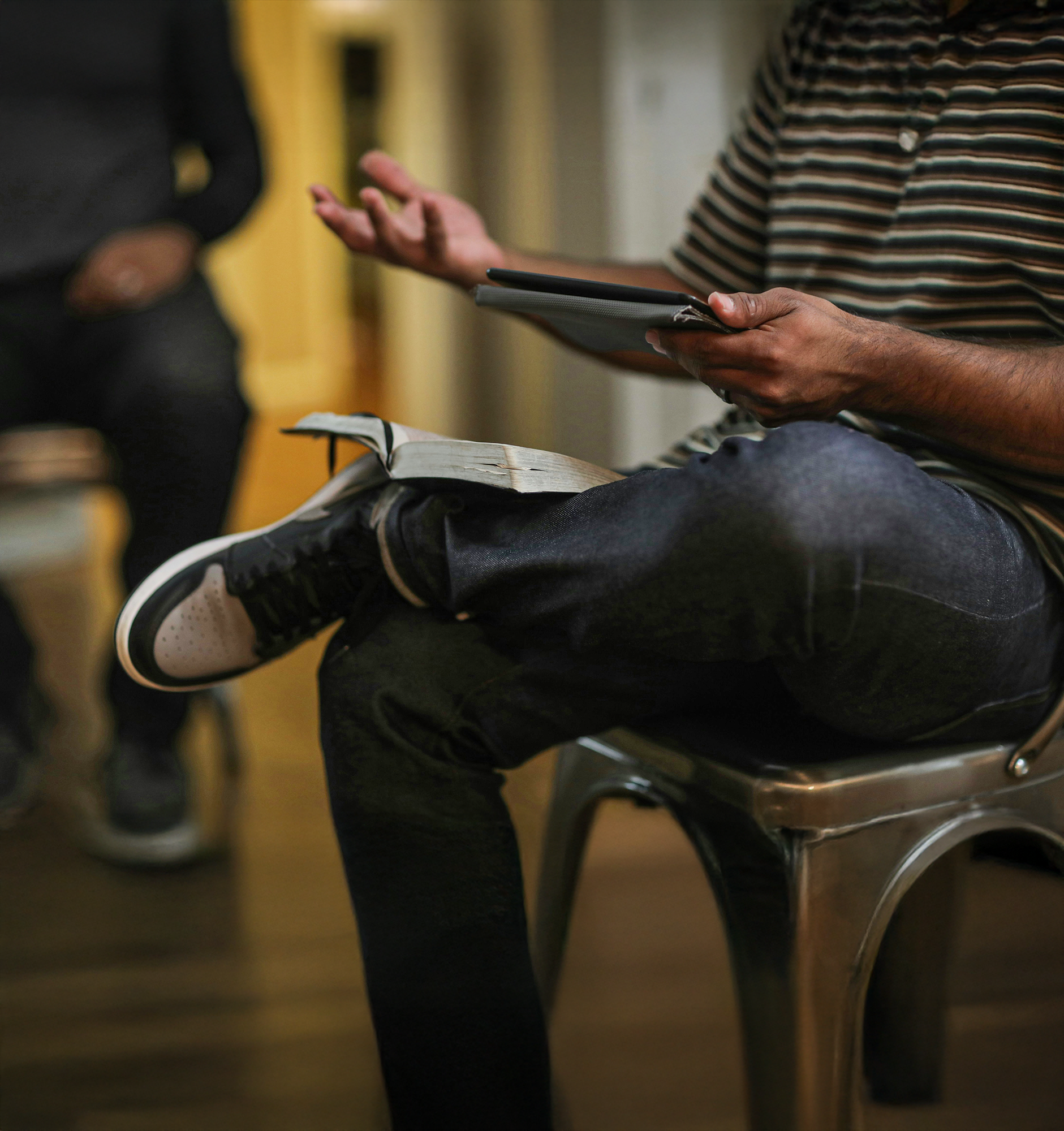 Person sitting cross-legged on a metal chair holding a tablet with an open book on their lap.