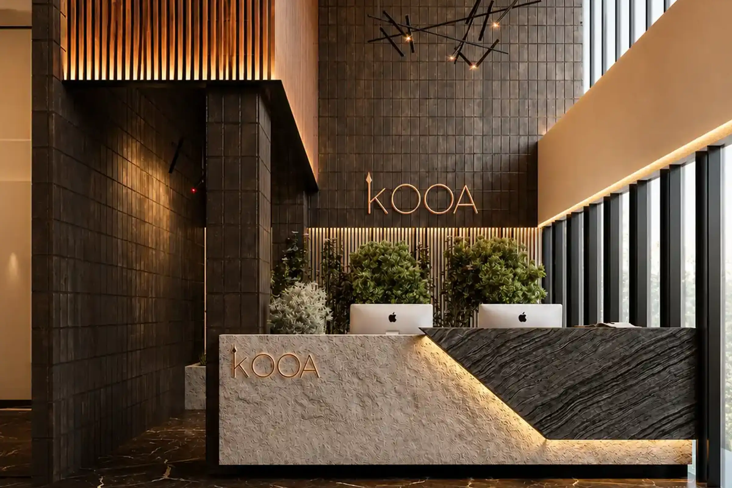 Modern lobby reception desk with KOAA logo, two iMacs, dark tiled walls, plants, and tall windows.