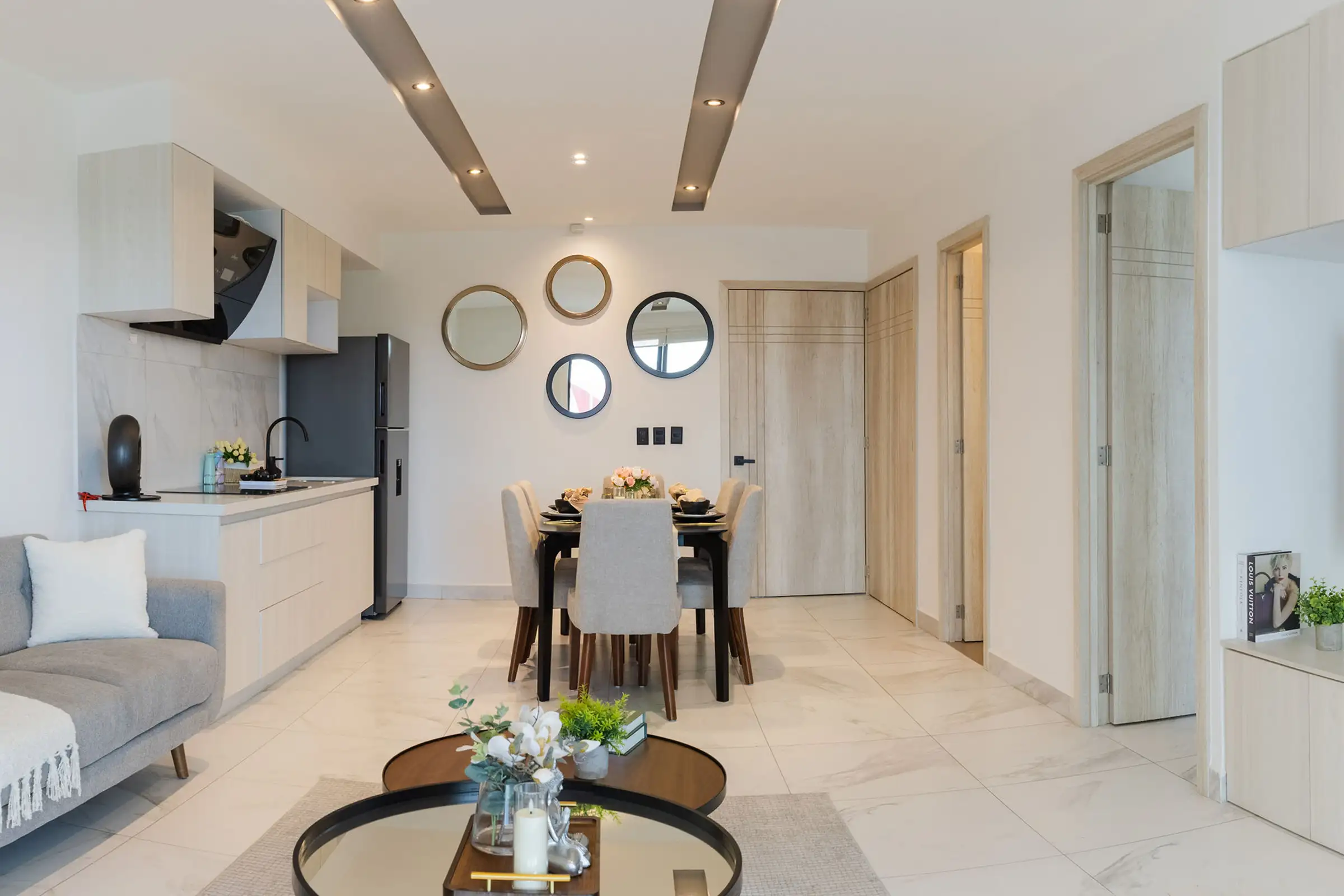 Modern open living area with kitchen, dining table, round mirrors, and a gray sofa with a white pillow.