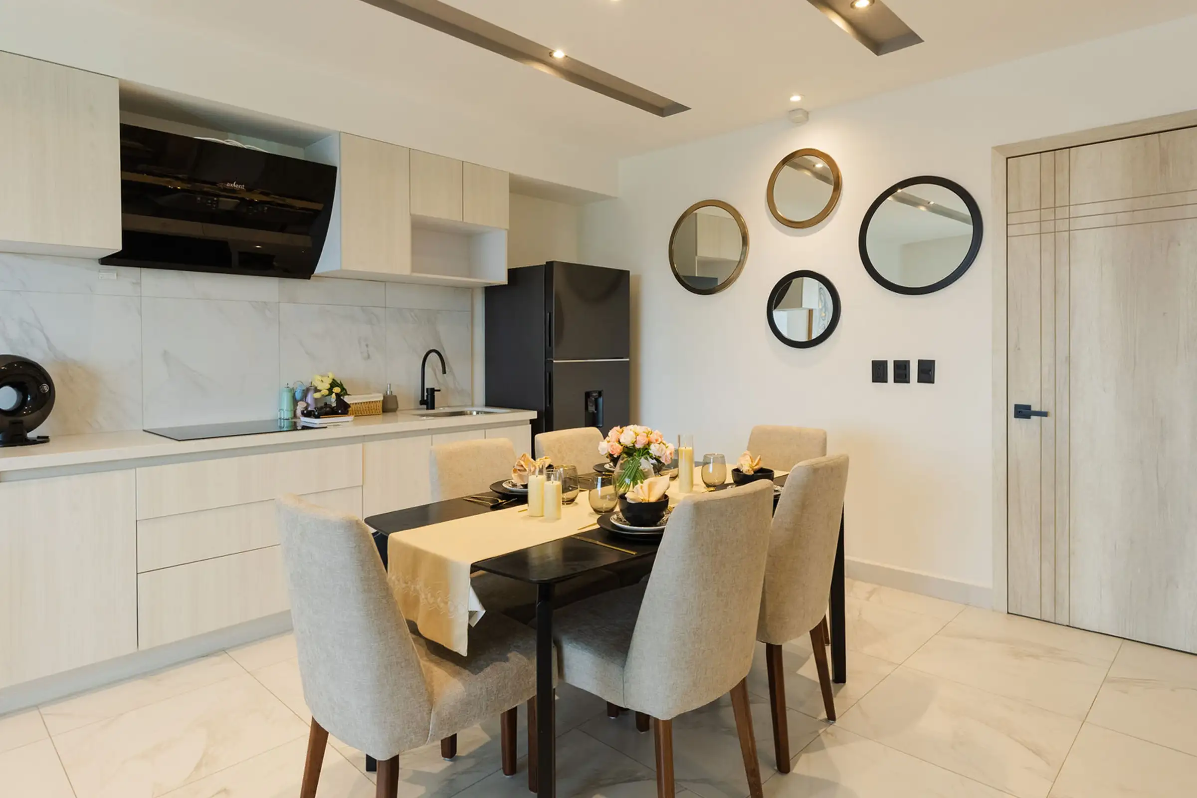 Modern dining area with six beige chairs, a black table, flower centerpiece, and a kitchen with light cabinets.