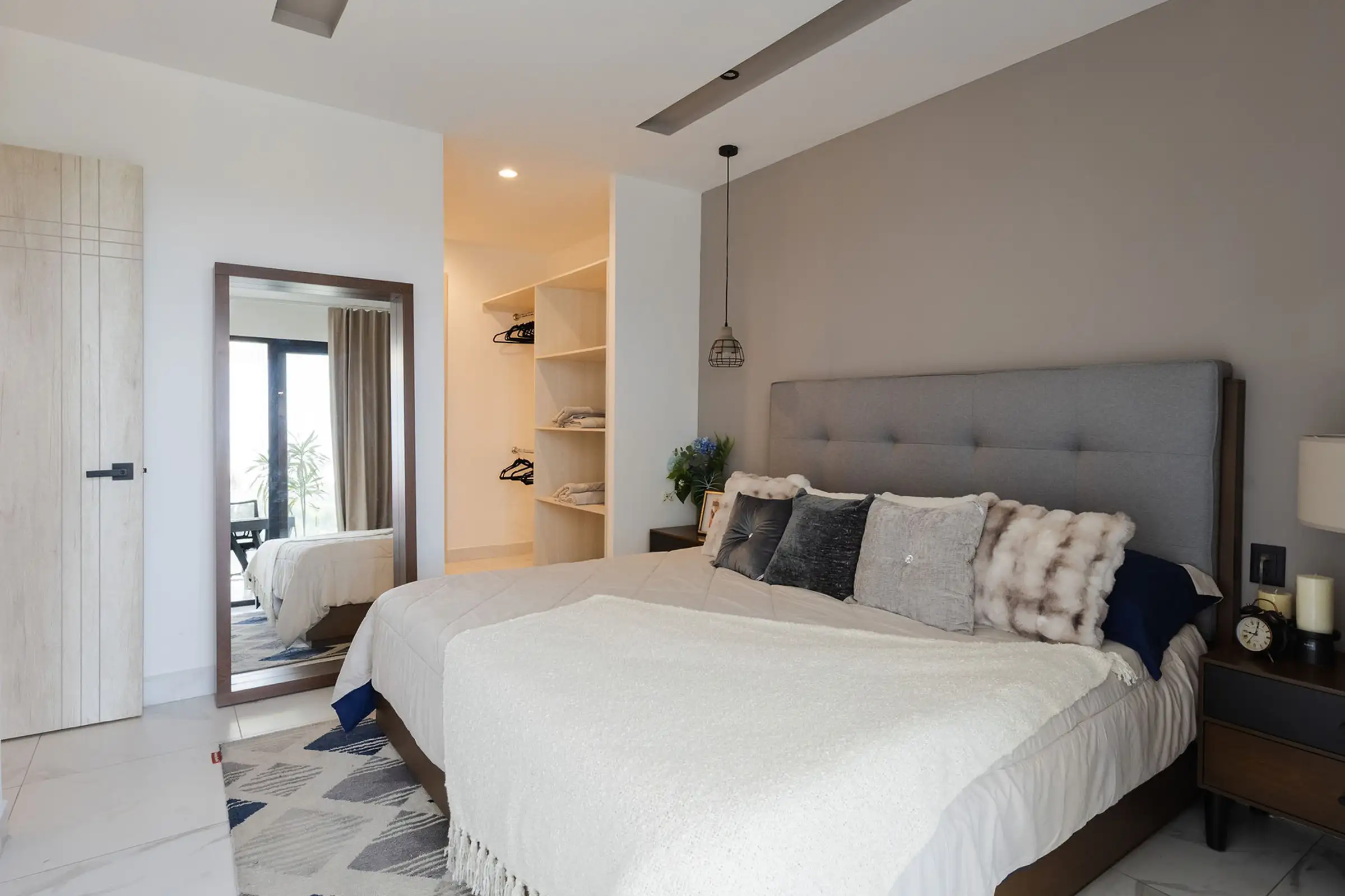 Modern bedroom with gray headboard bed, white blanket, decorative pillows, wooden door, and a standing mirror.