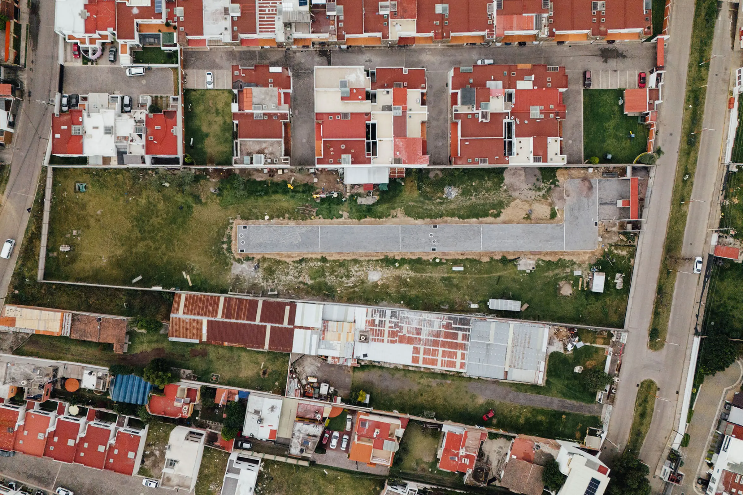 Aerial view of a residential neighborhood with rows of houses having orange and grey roofs, a large vacant construction site in the middle with construction vehicles and equipment, surrounded by streets with parked cars.